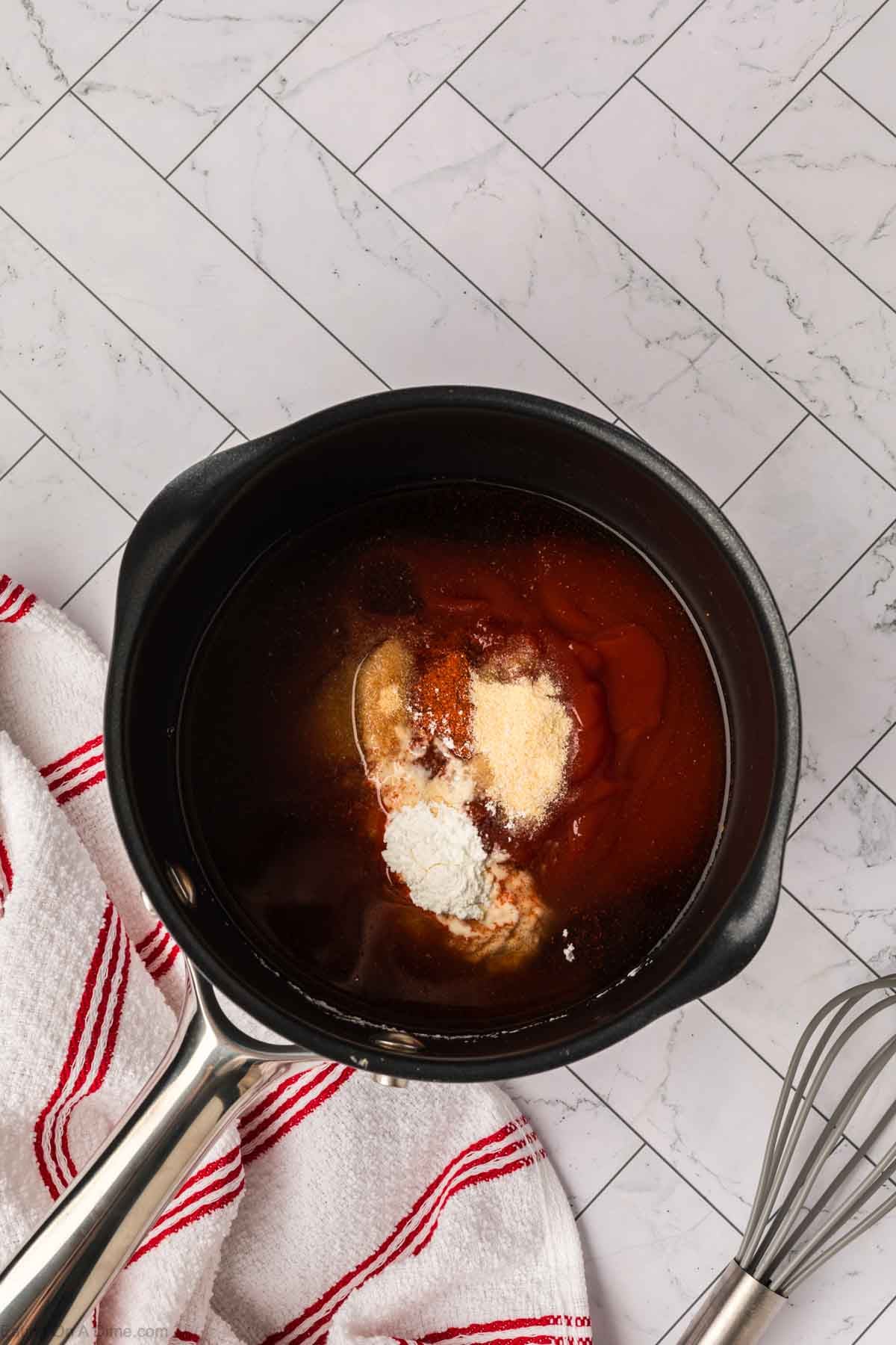A black saucepan with various spices, Arby’s Sauce, ketchup, and seasonings sits on a white tiled countertop beside a white towel with red stripes and a metal whisk.