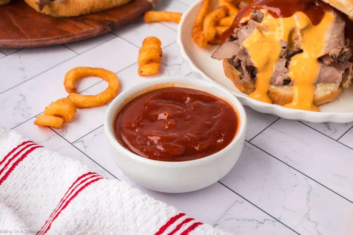 A small white bowl filled with Arby’s Sauce sits on a white tiled surface, with curly fries, a roast beef sandwich topped with cheese sauce, and a red-striped towel nearby.