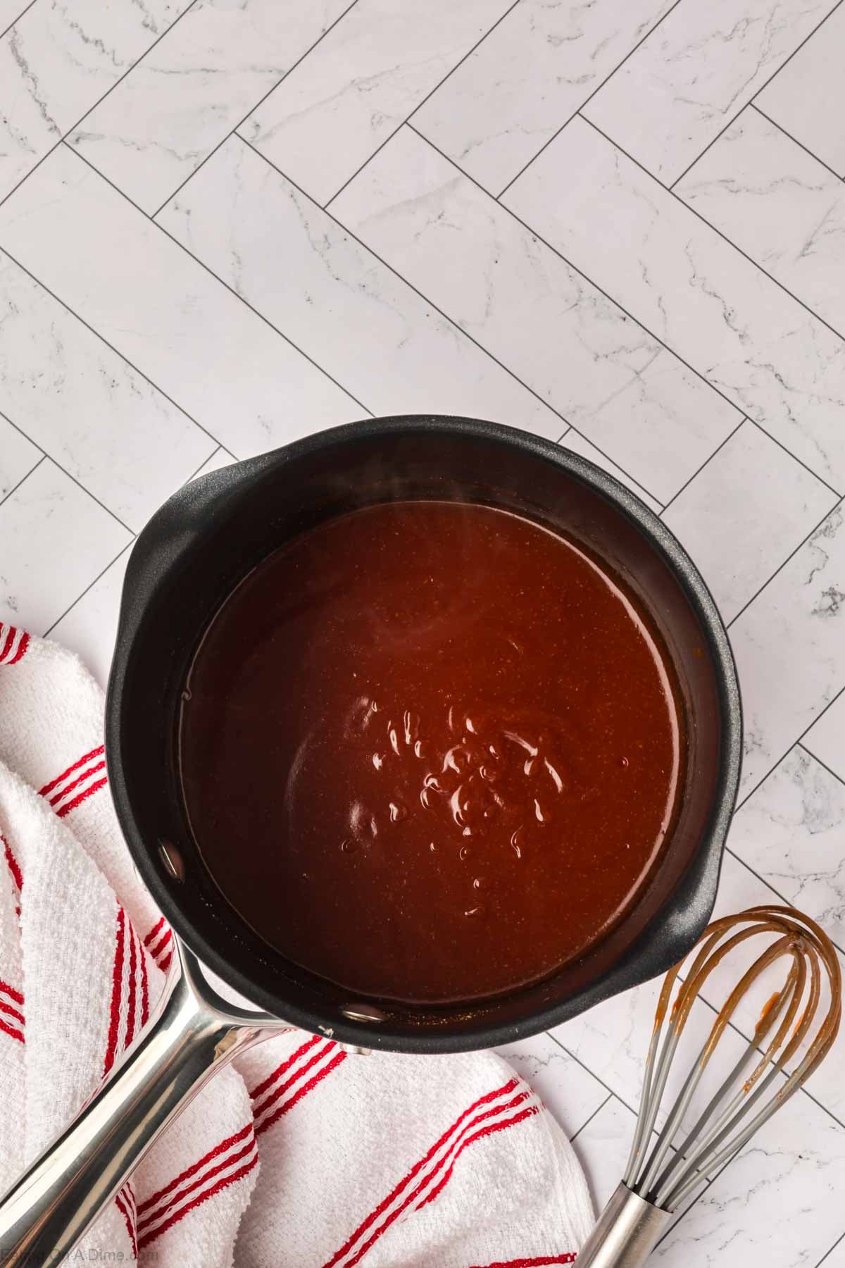 A saucepan filled with glossy brown chocolate sauce sits on a white striped towel beside a metal whisk, evoking the smooth richness of an Arby’s Sauce recipe, on a white tile countertop.