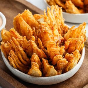 A bowl of crispy, golden-brown Bloomin Onion petals seasoned with spices, arranged closely together and served on a wooden tray.