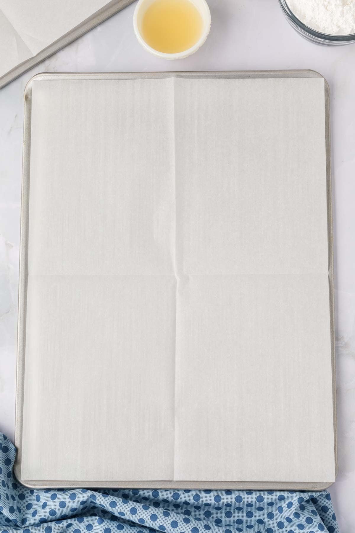 A baking sheet lined with parchment paper sits on a marble surface, ready for a batch of blueberry cookies. Nearby are a small bowl of yellow liquid, a bowl of white powder, and a blue polka dot cloth.