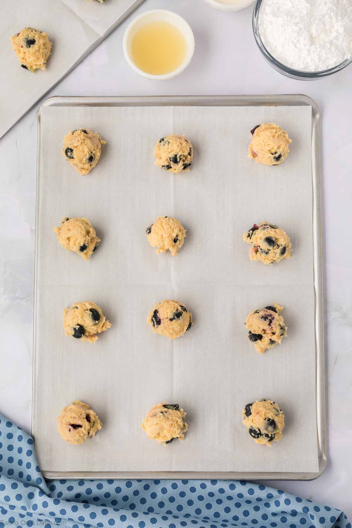 A baking sheet lined with parchment paper holds twelve scoops of unbaked Blueberry Cookies dough. Nearby are a bowl of flour, a small bowl of liquid, and a blue polka-dot cloth.