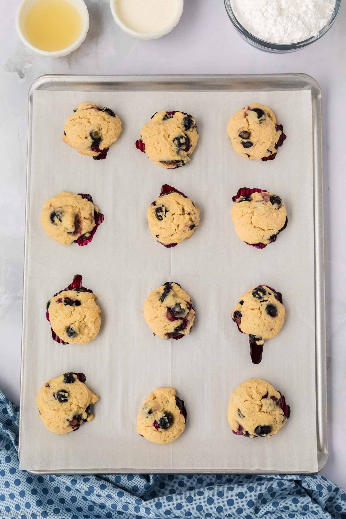 A baking sheet lined with parchment paper holds twelve freshly baked blueberry cookies. Some of the blueberries have burst, leaving purple stains. Bowls with ingredients and a blue polka-dot cloth hint at more blueberry cookies to come.
