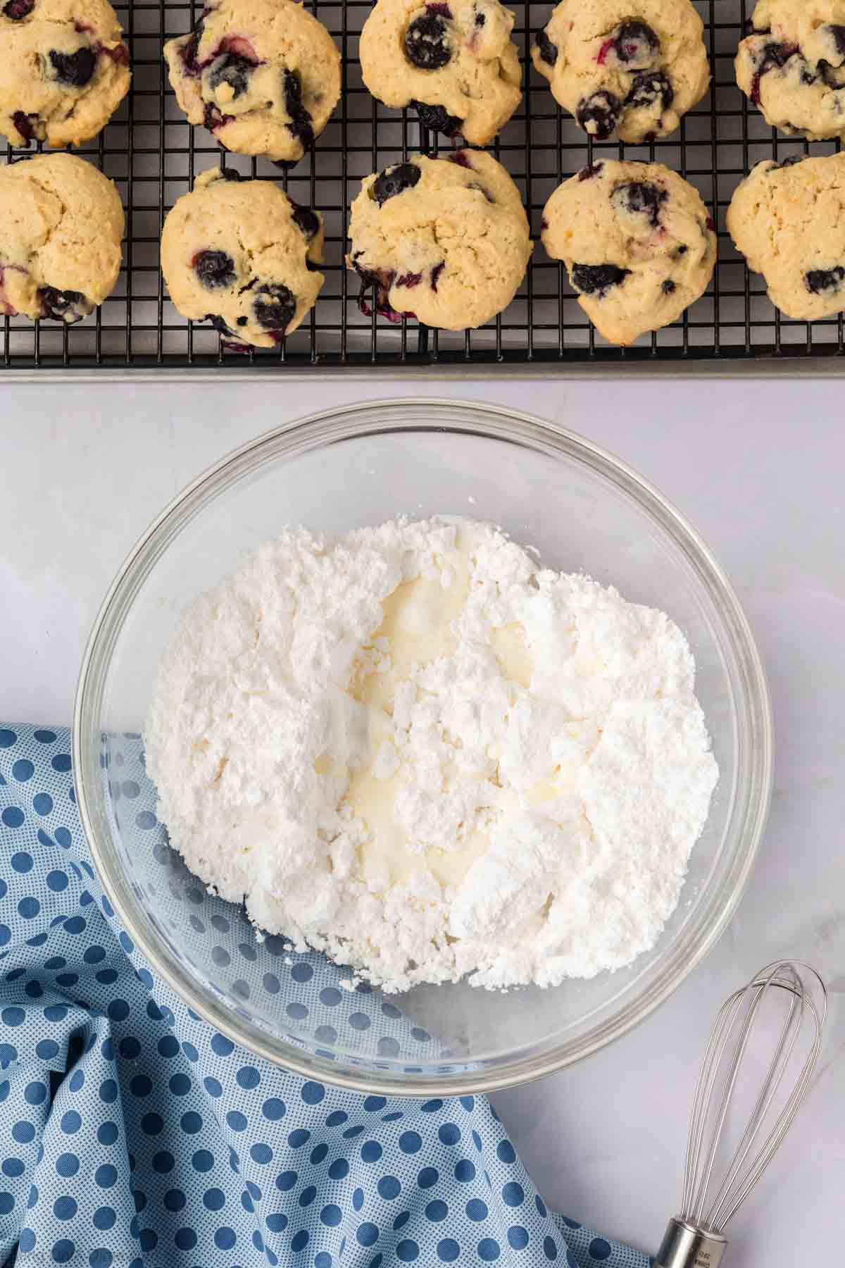 A glass bowl with powdered sugar and liquid sits on a countertop next to a metal whisk and a blue polka dot cloth. In the background, freshly baked blueberry cookies cool on a wire rack.