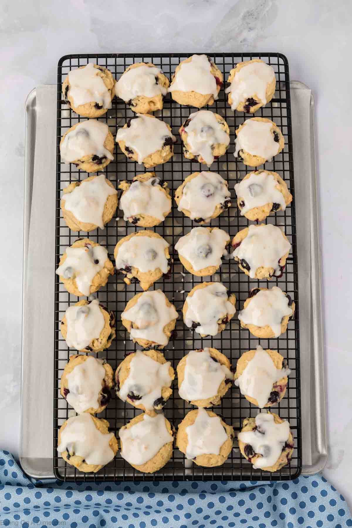 A cooling rack filled with rows of blueberry cookies, their glazed surfaces revealing pockets of juicy berries, set on a baking sheet with a blue and white polka dot cloth partially underneath.