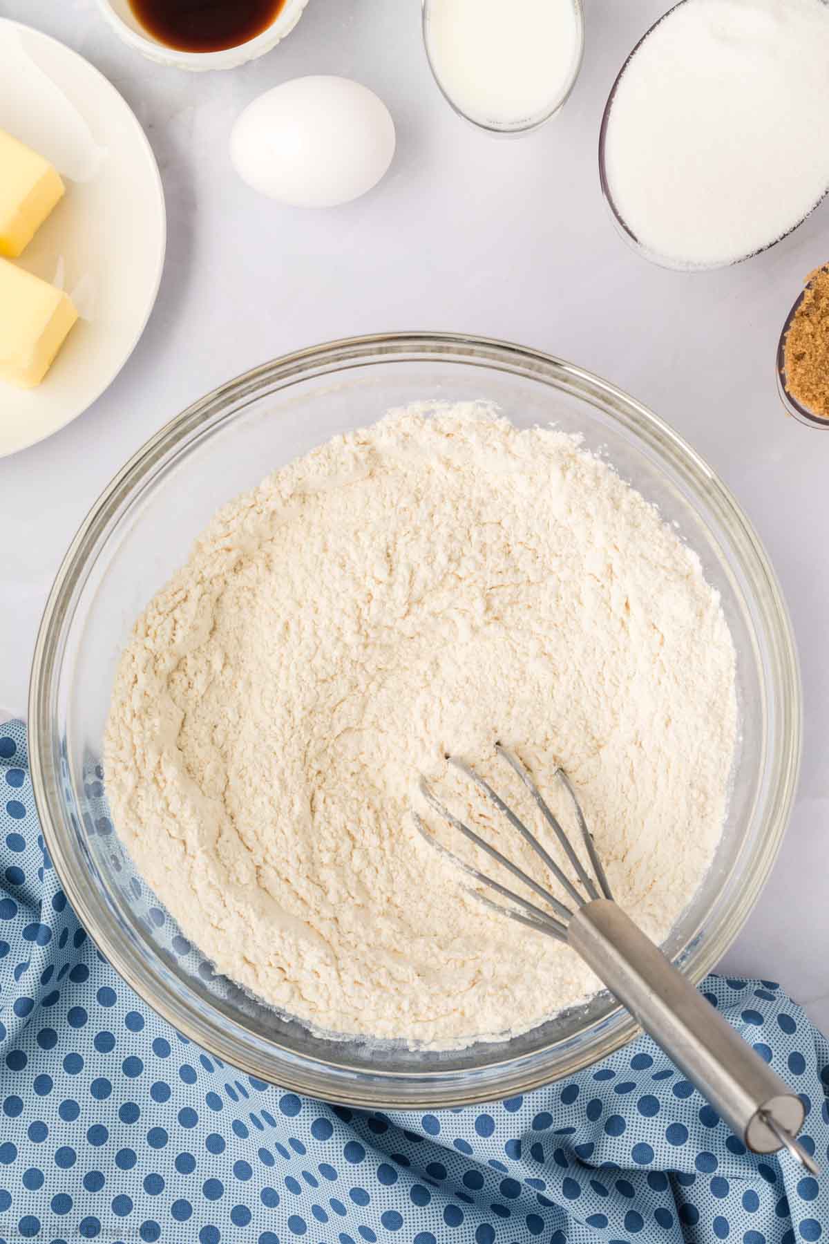 A glass bowl with flour mixture and a whisk, surrounded by ingredients for Blueberry Cookies—butter, an egg, milk, vanilla extract, white sugar, and brown sugar—on a white surface with a blue polka dot cloth.
