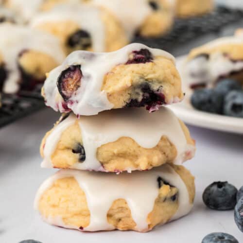 Three Blueberry Cookies topped with white icing are stacked on a white surface, surrounded by fresh blueberries and more cookies in the background.