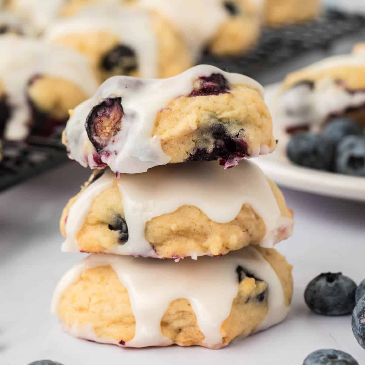 Three Blueberry Cookies topped with white icing are stacked on a white surface, surrounded by fresh blueberries and more cookies in the background.