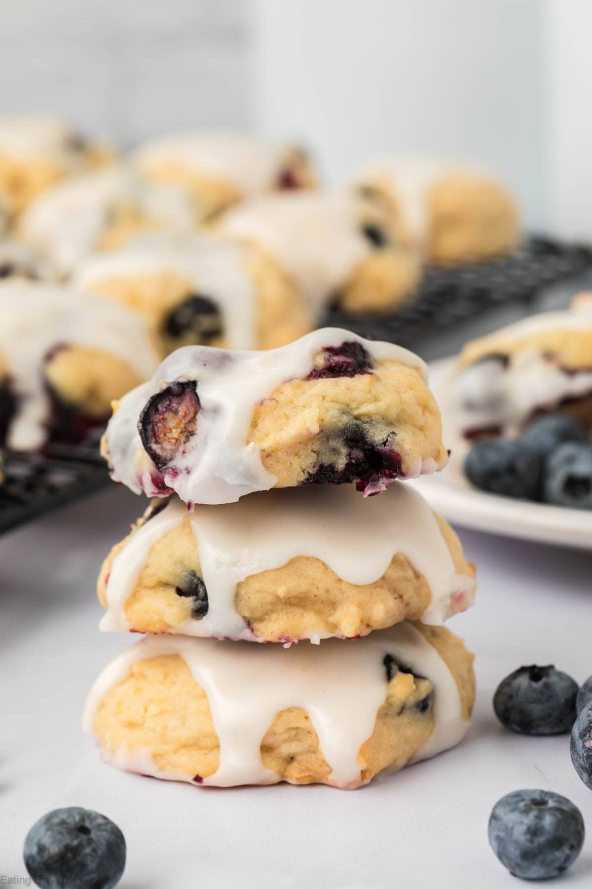 Three blueberry cookies with white icing are stacked on top of each other, surrounded by fresh blueberries. More blueberry cookies rest in the background on a cooling rack and plate, creating an inviting display.