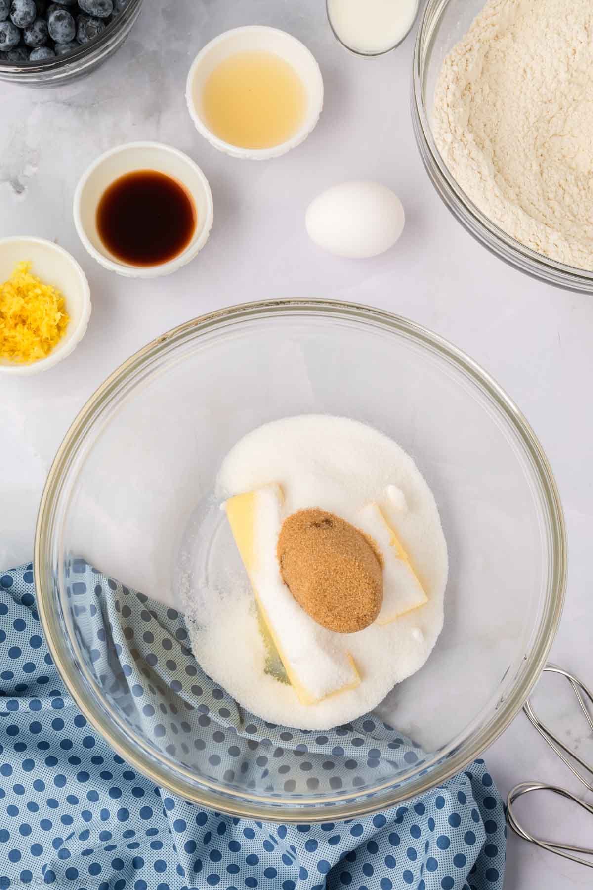 A glass bowl with sugar, brown sugar, and butter sits on a counter surrounded by ingredients for Blueberry Cookies: lemon zest, vanilla extract, lemon juice, an egg, flour, blueberries, plus a blue polka-dot cloth and hand mixer beaters.