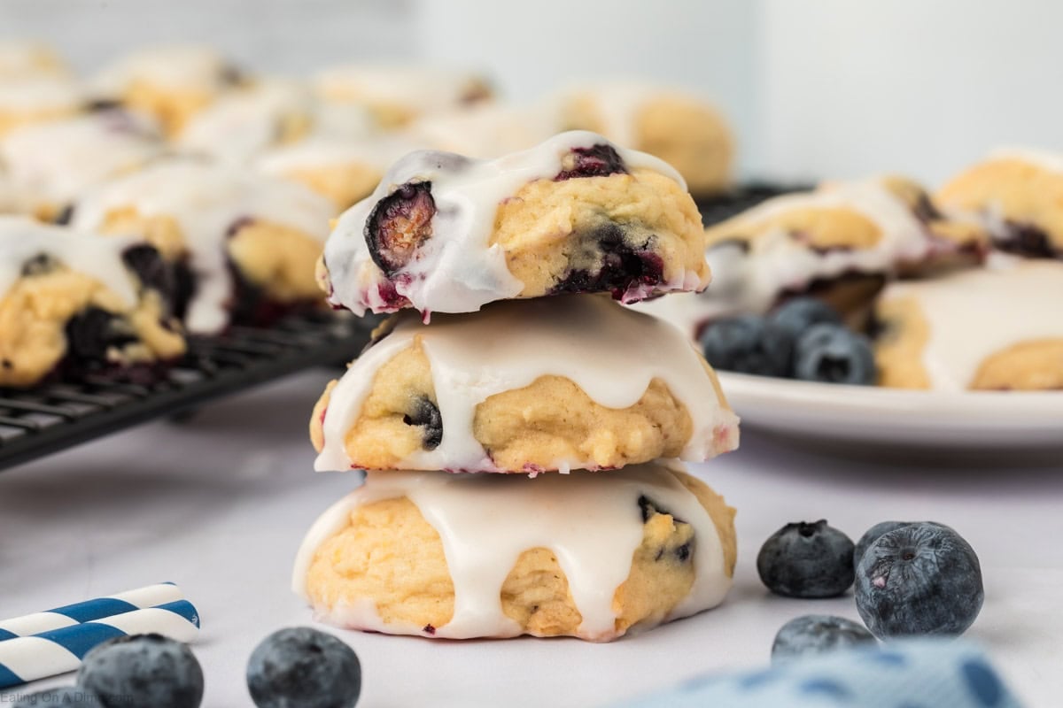 Three Blueberry Cookies with white icing are stacked on a table, surrounded by fresh blueberries. More Blueberry Cookies and a plate appear in the blurred background, evoking a fresh, homemade dessert setting.