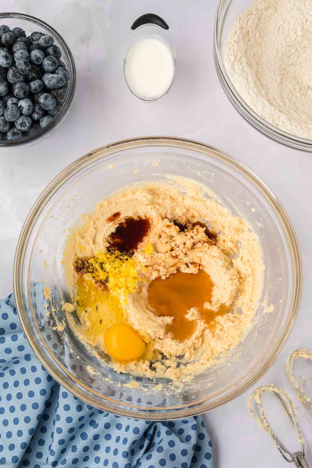 A glass bowl with creamed butter, egg, vanilla, lemon zest, and sugar—ingredients ready for delicious Blueberry Cookies. Nearby are a bowl of blueberries, flour, a cup of milk, a blue polka dot cloth, and a hand mixer.