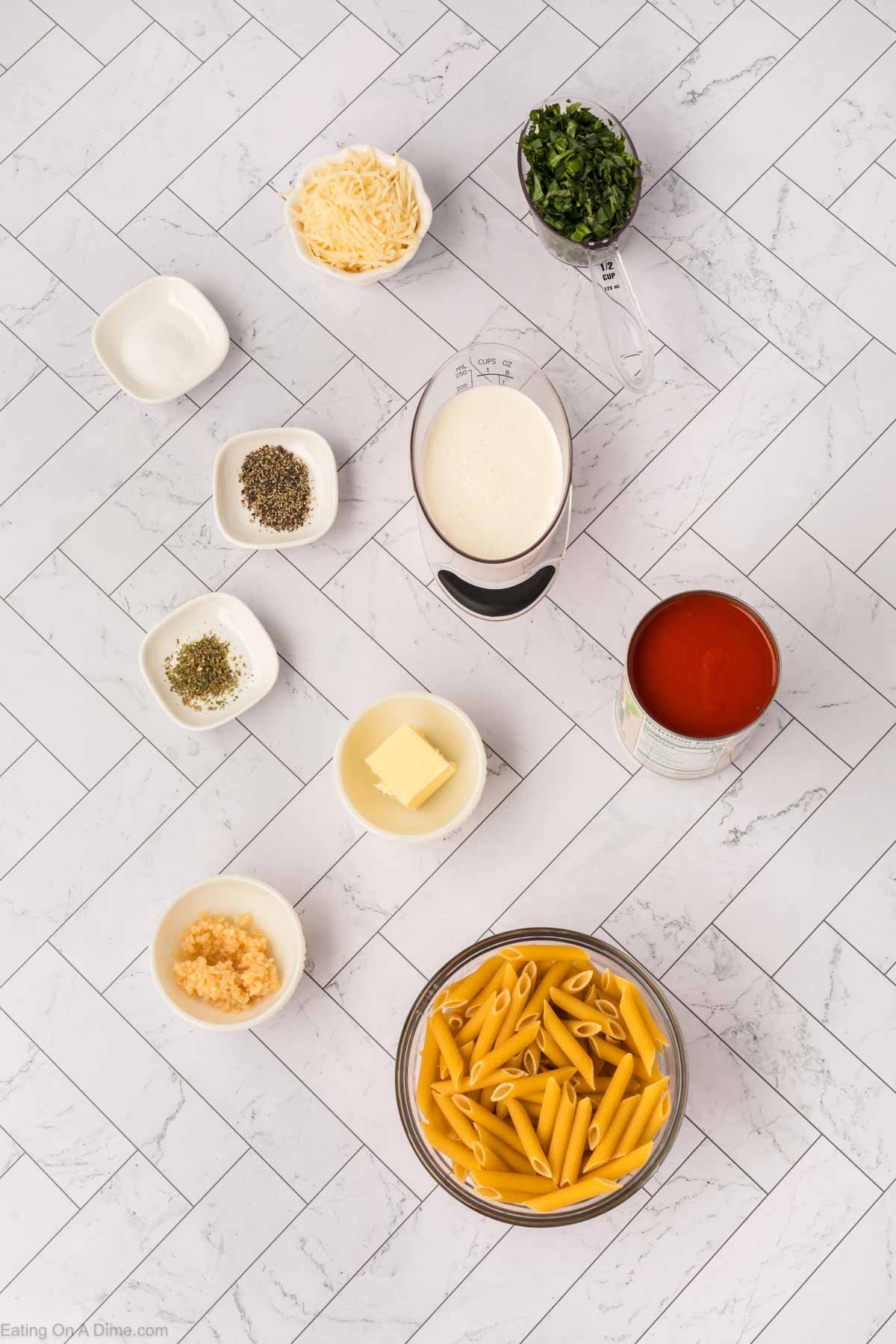 Overhead view of Tomato Basil Pasta ingredients—dry penne, butter, minced garlic, Italian seasonings, herbs, milk, shredded cheese, chopped parsley, tomato sauce, and salt—artfully arranged on a white tile surface.