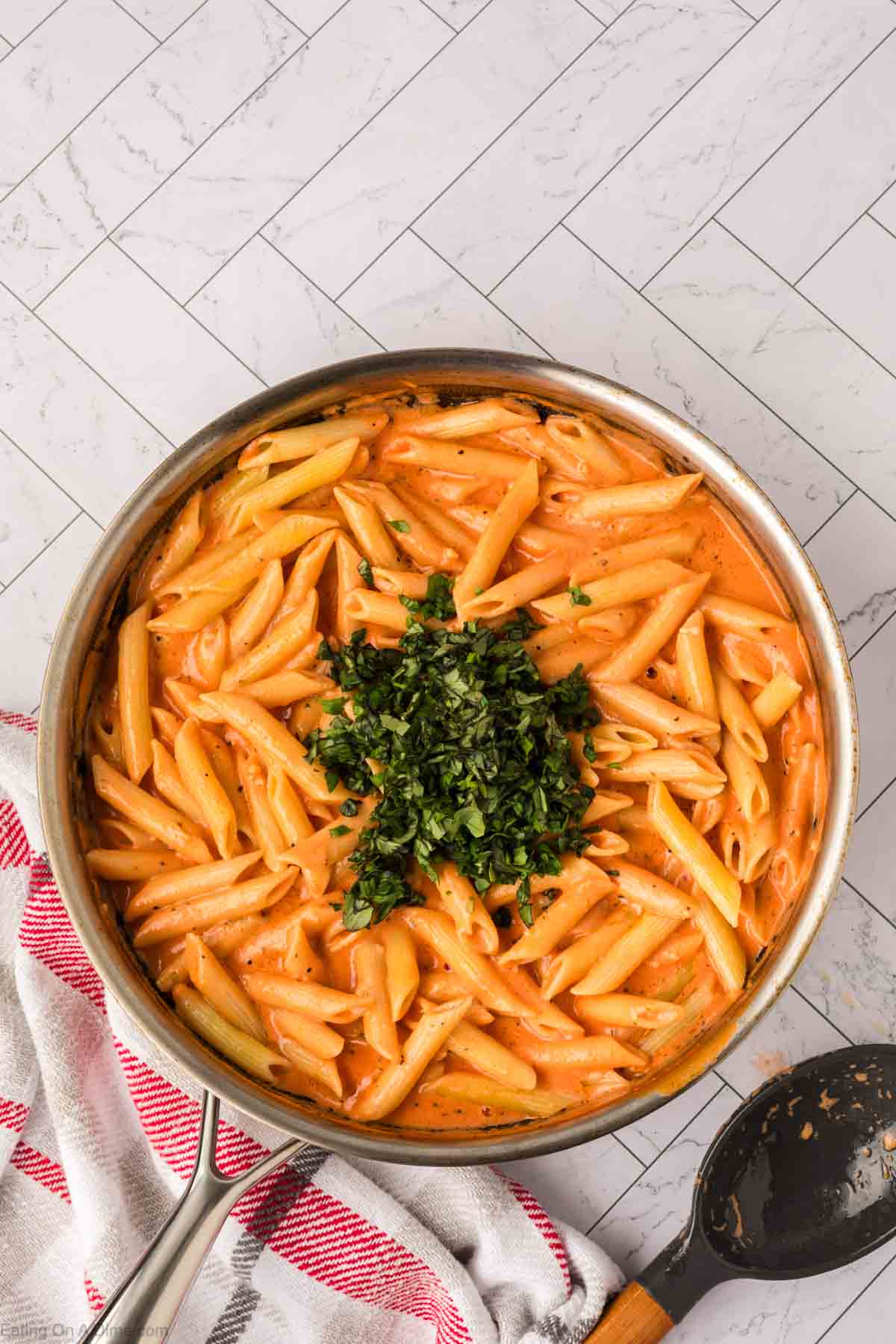 A skillet filled with creamy pasta, penne in a rich tomato basil sauce, topped with freshly chopped herbs. A red-and-white kitchen towel and a black serving spoon are beside the skillet on a light, tiled countertop.