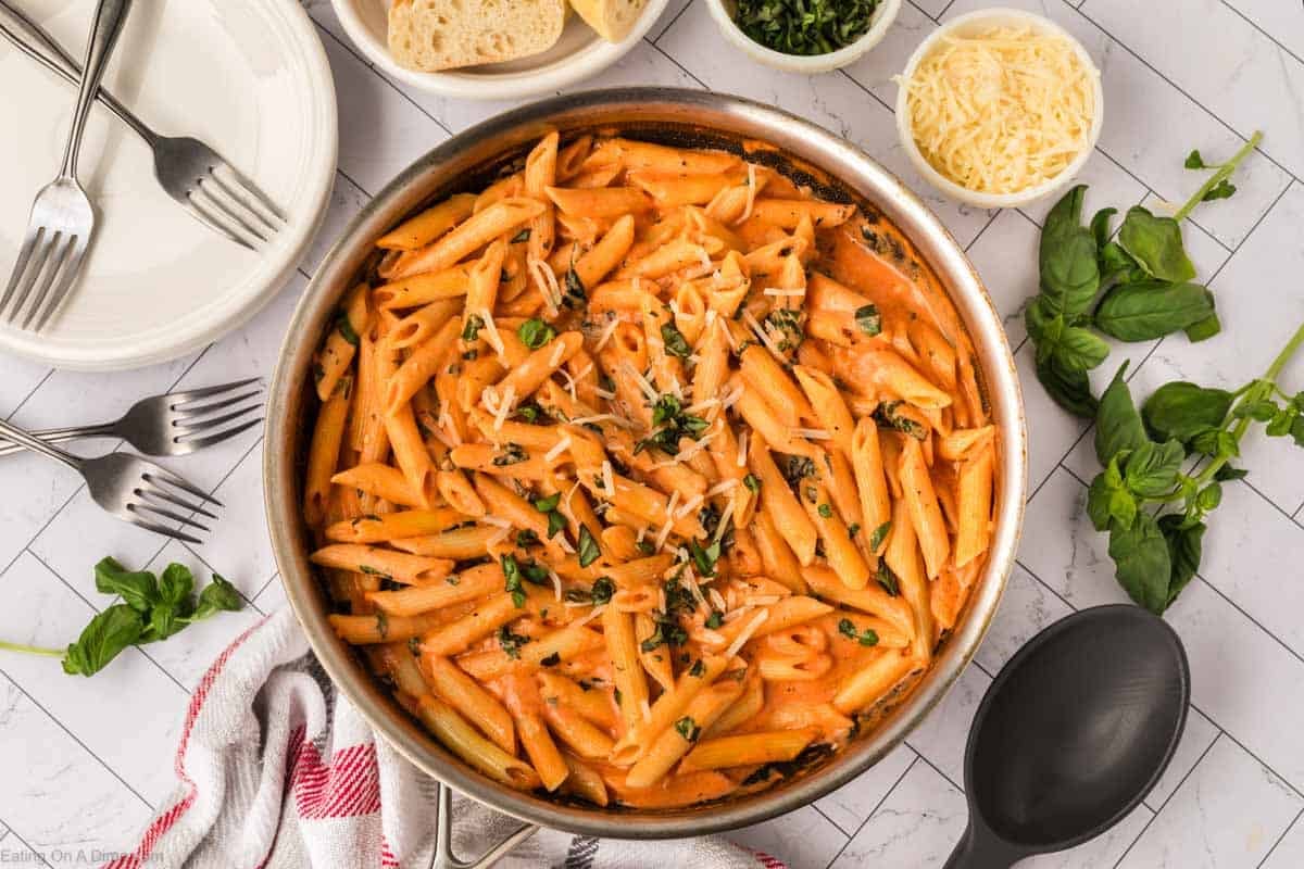 A skillet filled with creamy tomato pasta, topped with shredded cheese and chopped basil, surrounded by plates, forks, bread, grated cheese, basil leaves, and a serving spoon on a white surface.