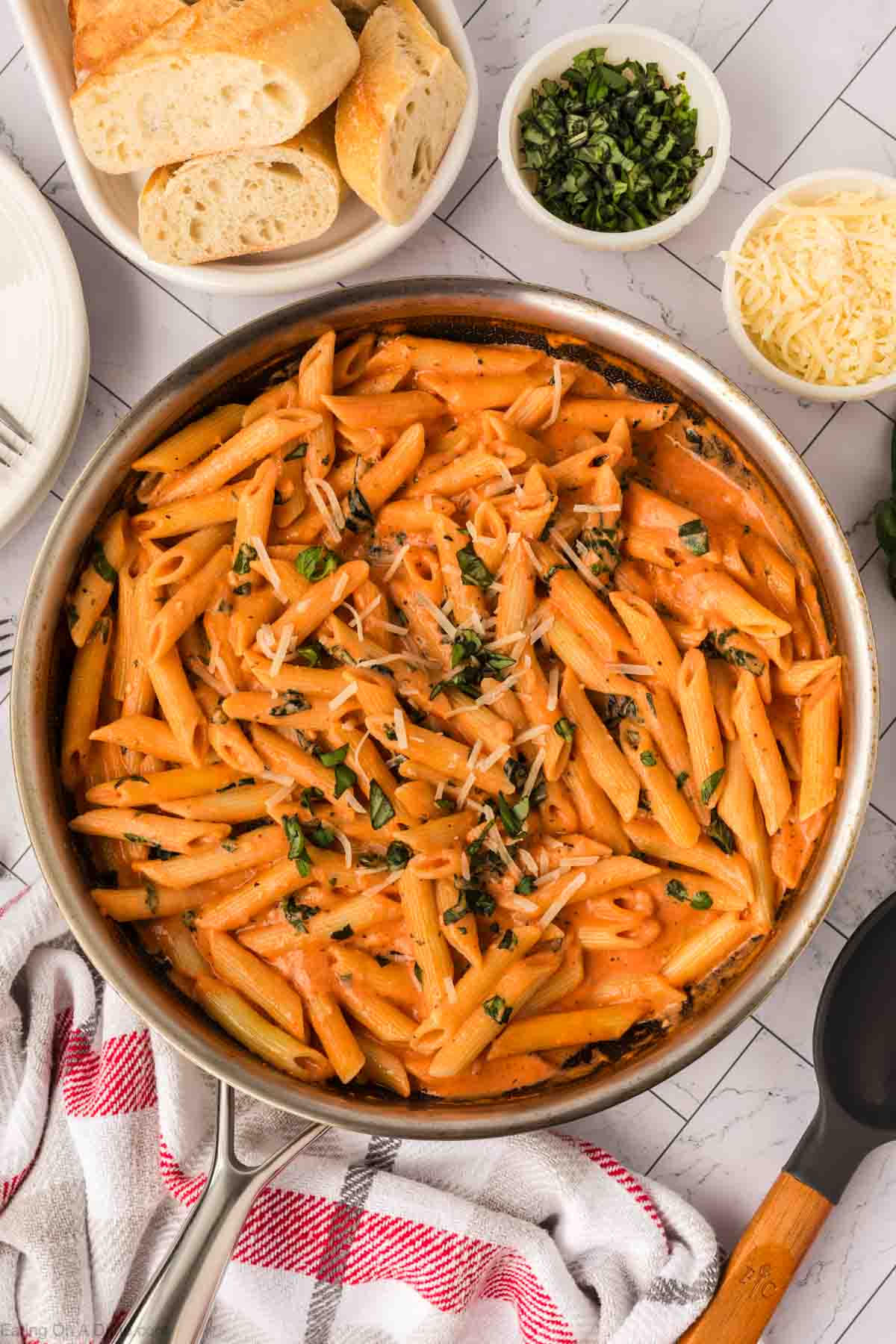A skillet filled with Creamy Tomato Basil Pasta, topped with chopped basil and grated cheese. Surrounding the skillet are a plate of sliced bread, a bowl of shredded cheese, and fresh herbs on a white surface.