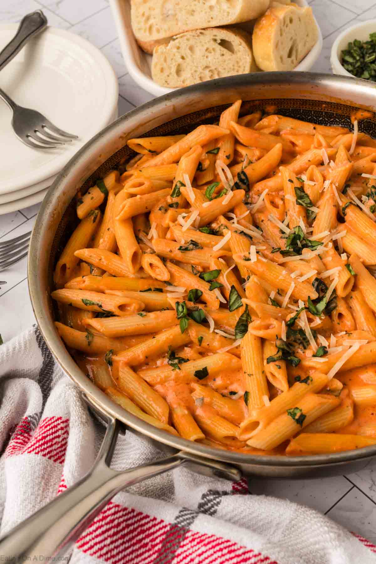 A skillet filled with creamy tomato pasta, garnished with shredded cheese and fresh basil, sits on a red and white towel. Slices of bread and plates with forks are in the background.