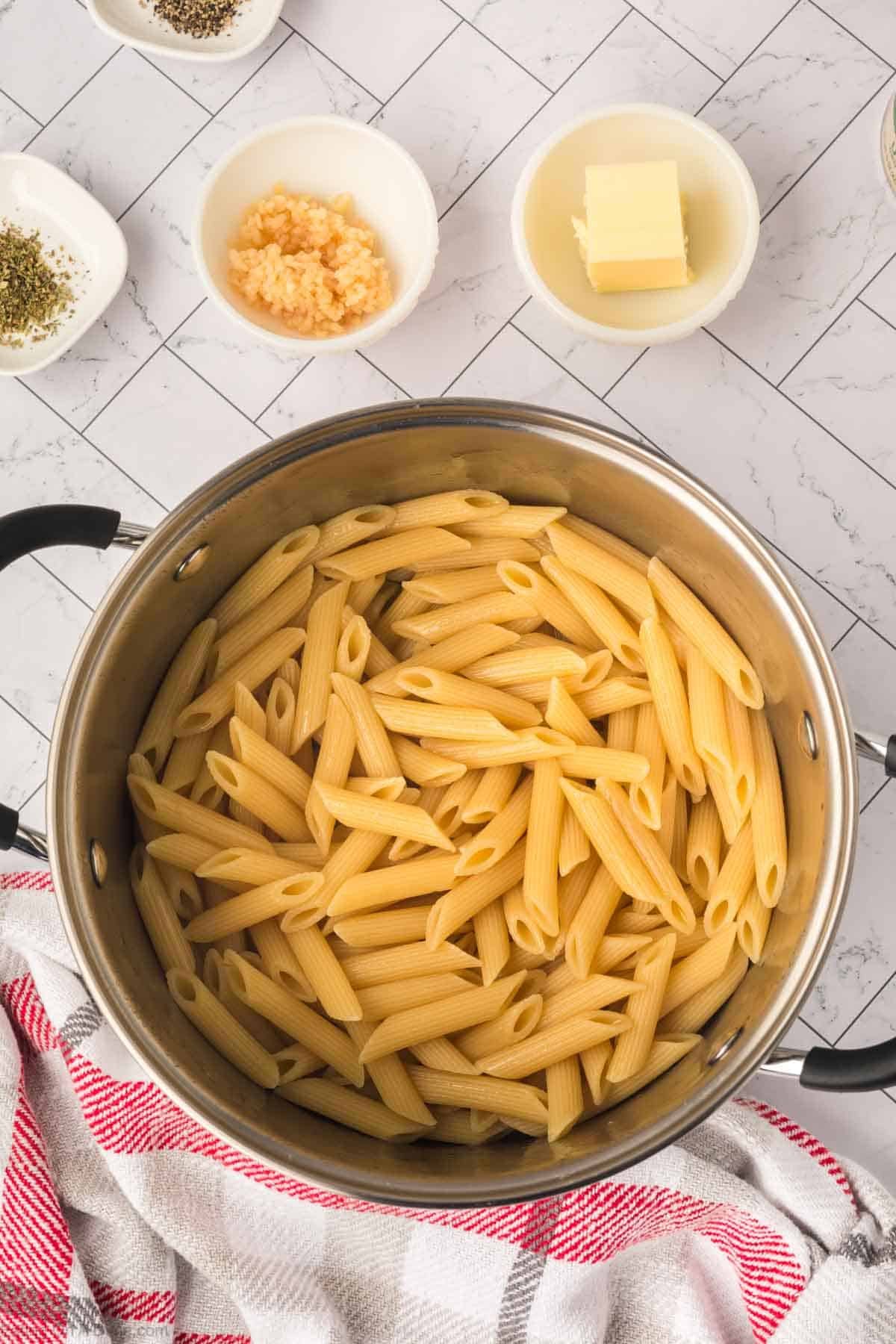 A pot of cooked penne pasta sits on a countertop next to bowls containing minced garlic, butter, and seasonings—perfect for making creamy tomato pasta. A red and white kitchen towel is draped nearby.