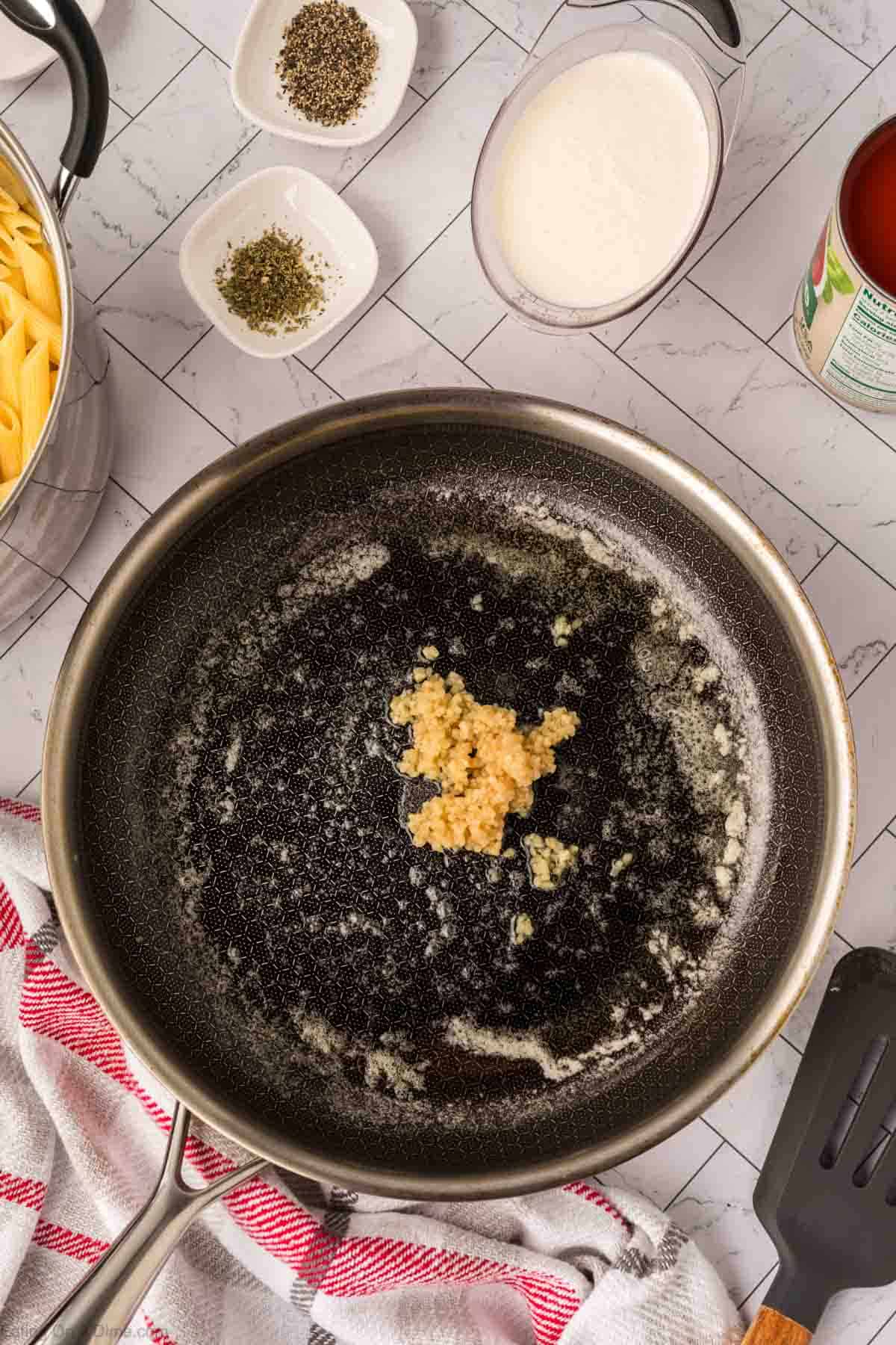 A skillet with melted butter and minced garlic cooks on a stovetop. Surrounding the pan are bowls of dried herbs, a pitcher of cream, a can of tomatoes, cooked pasta, and a red-striped towel—perfect for creamy tomato basil pasta.