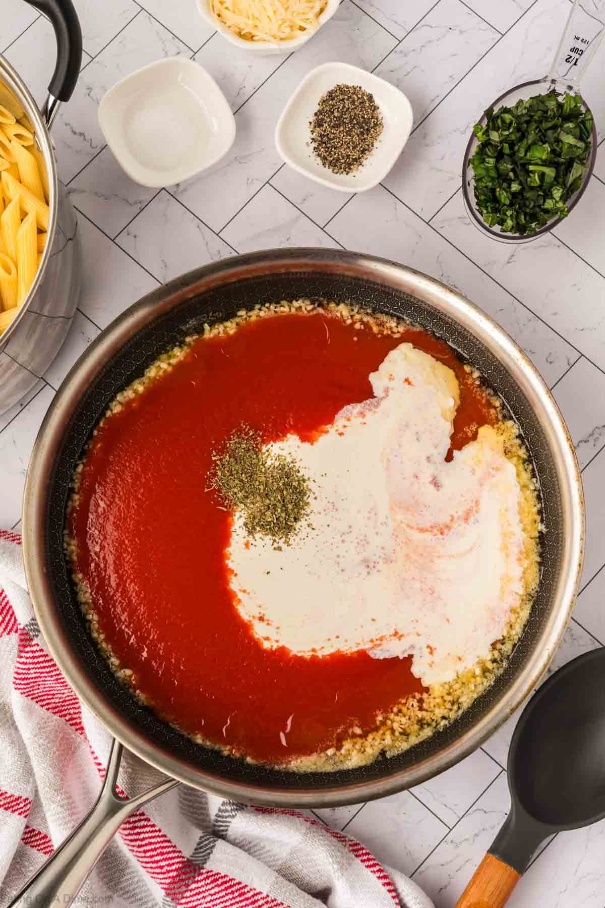 A skillet contains creamy tomato basil pasta sauce being mixed. Surrounding the skillet are bowls of chopped herbs, shredded cheese, pepper, penne pasta, and a towel on a white tiled countertop.