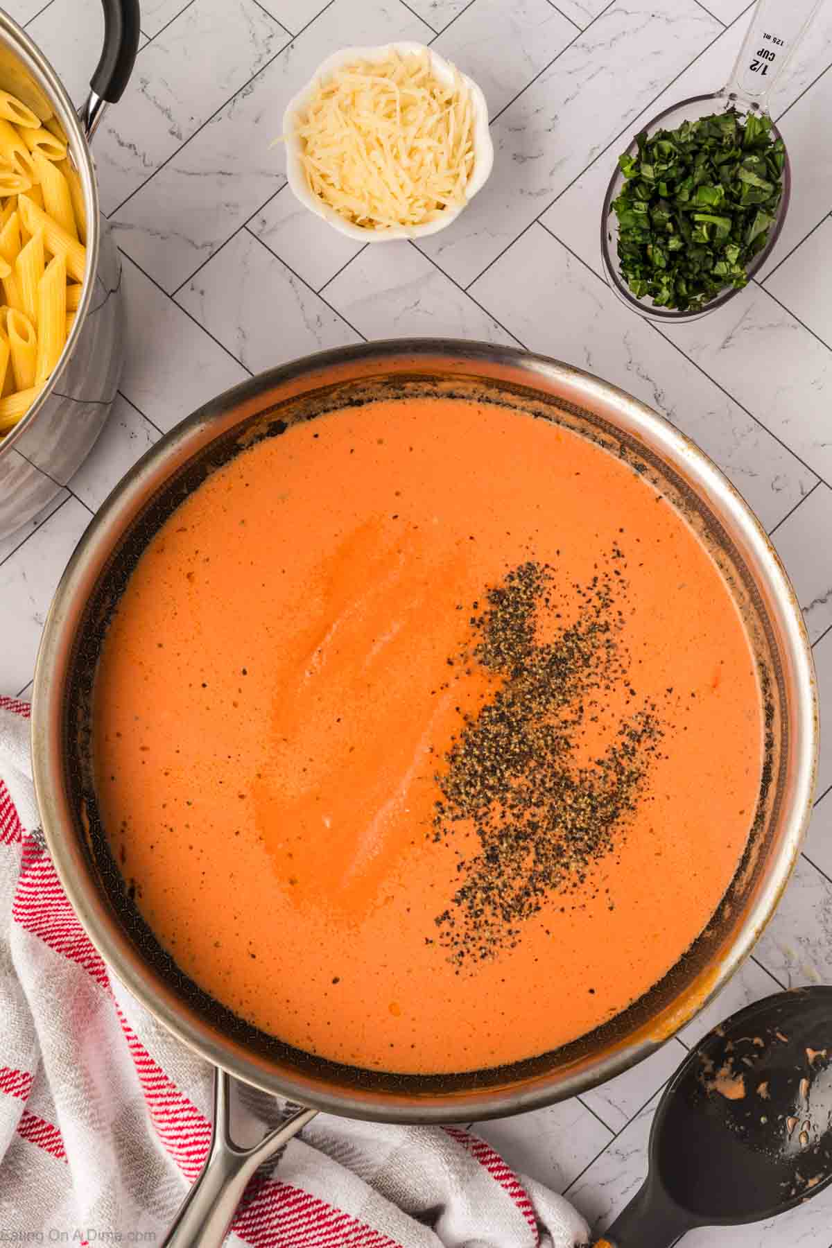 A skillet filled with creamy tomato sauce, perfect for Creamy Tomato Basil Pasta, topped with cracked black pepper and surrounded by a bowl of shredded cheese, chopped herbs, and a pot of cooked penne on the kitchen countertop.