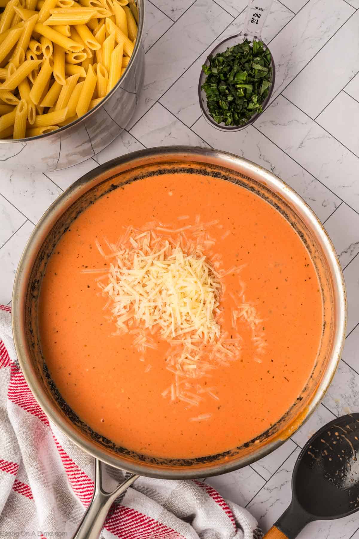 A pan of creamy tomato sauce topped with shredded cheese sits on a countertop. Nearby are a bowl of uncooked penne pasta, chopped basil in a measuring cup, and a towel—perfect for making Creamy Tomato Basil Pasta.
