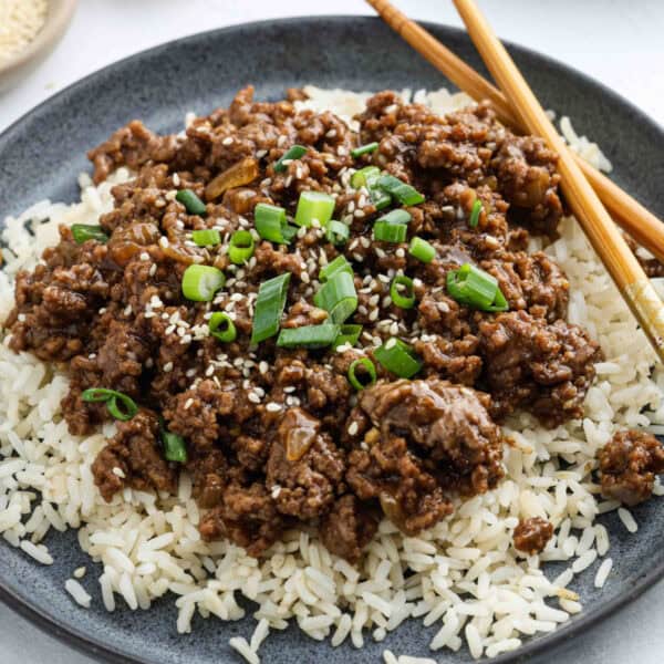 A plate of white rice topped with savory Korean Beef Bowls Recipe ground beef, garnished with chopped green onions and sesame seeds. Two wooden chopsticks rest on the side of the plate.