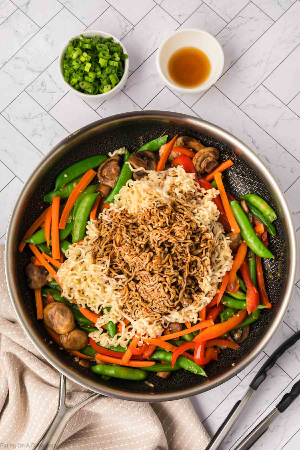 A skillet filled with cooked ramen noodles, sliced carrots, snap peas, mushrooms, and red bell peppers is transformed into a flavorful ramen stir fry with sauce poured on top. Nearby are bowls of chopped green onions and extra sauce on a tiled surface.