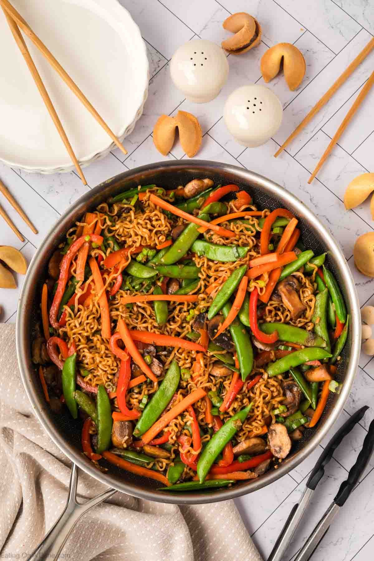 A skillet filled with savory ramen stir fry—stir-fried noodles, snap peas, red bell peppers, and mushrooms. Surrounding the skillet are chopsticks, fortune cookies, salt and pepper shakers, and an empty white plate.