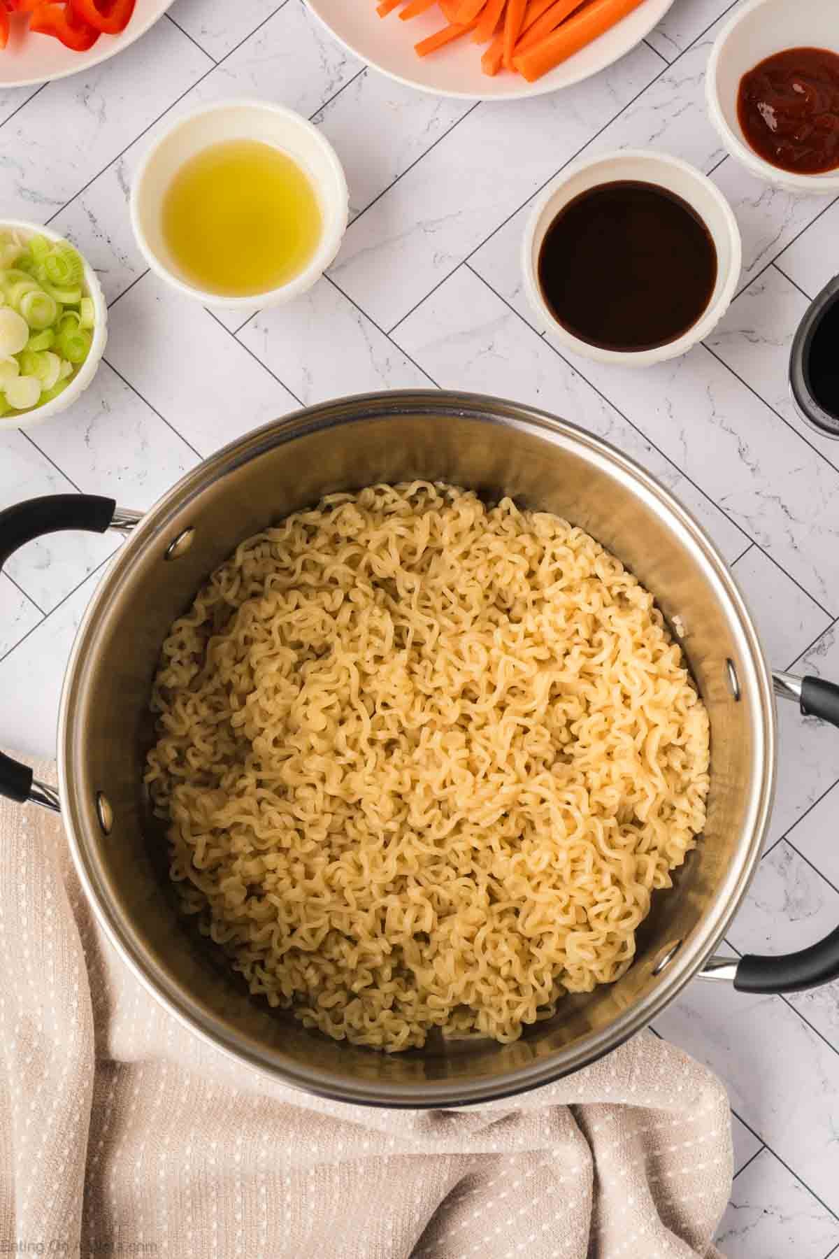 A pot of cooked ramen sits on a countertop, ready for a delicious ramen stir fry. Surrounding it are small bowls with sliced green onions, carrot sticks, sauce, and oil. A beige and white striped cloth is partially visible beside the pot.