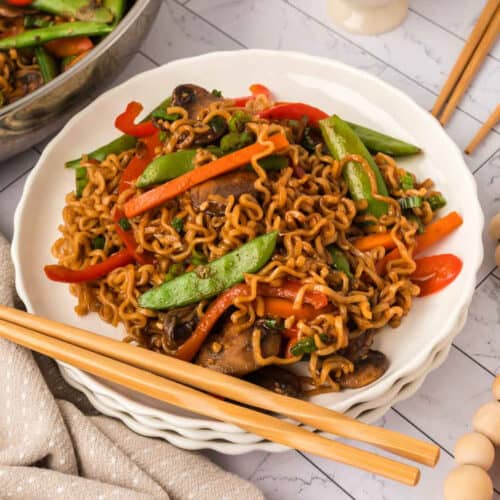 A plate of Ramen Stir Fry noodles with snap peas, red bell peppers, mushrooms, and savory sauce, served with chopsticks on a white plate.