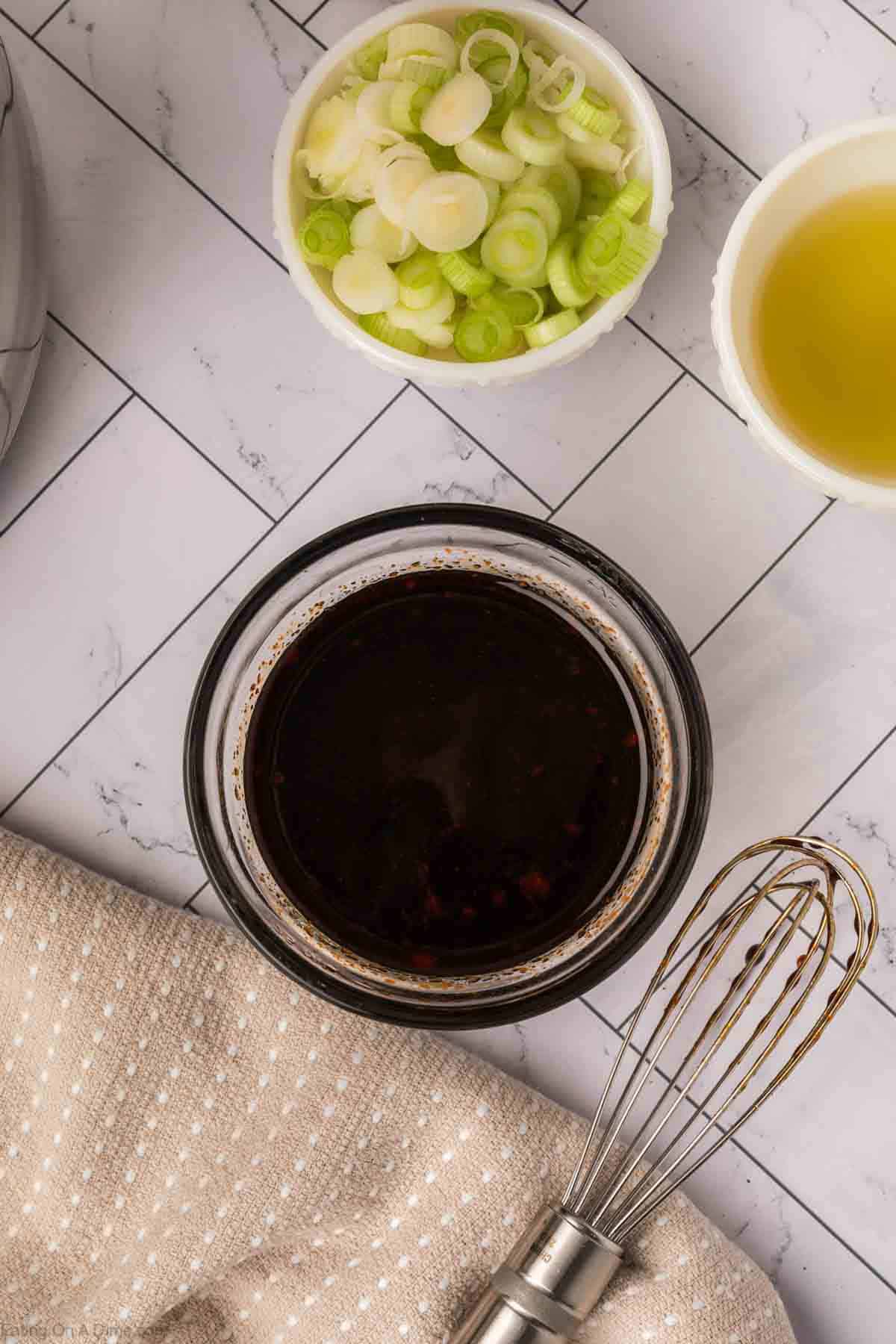 A bowl of dark soy-based sauce sits next to chopped green onions, a bowl of olive oil, and a metal whisk on a white-tiled surface—perfect ingredients for a delicious ramen stir fry.