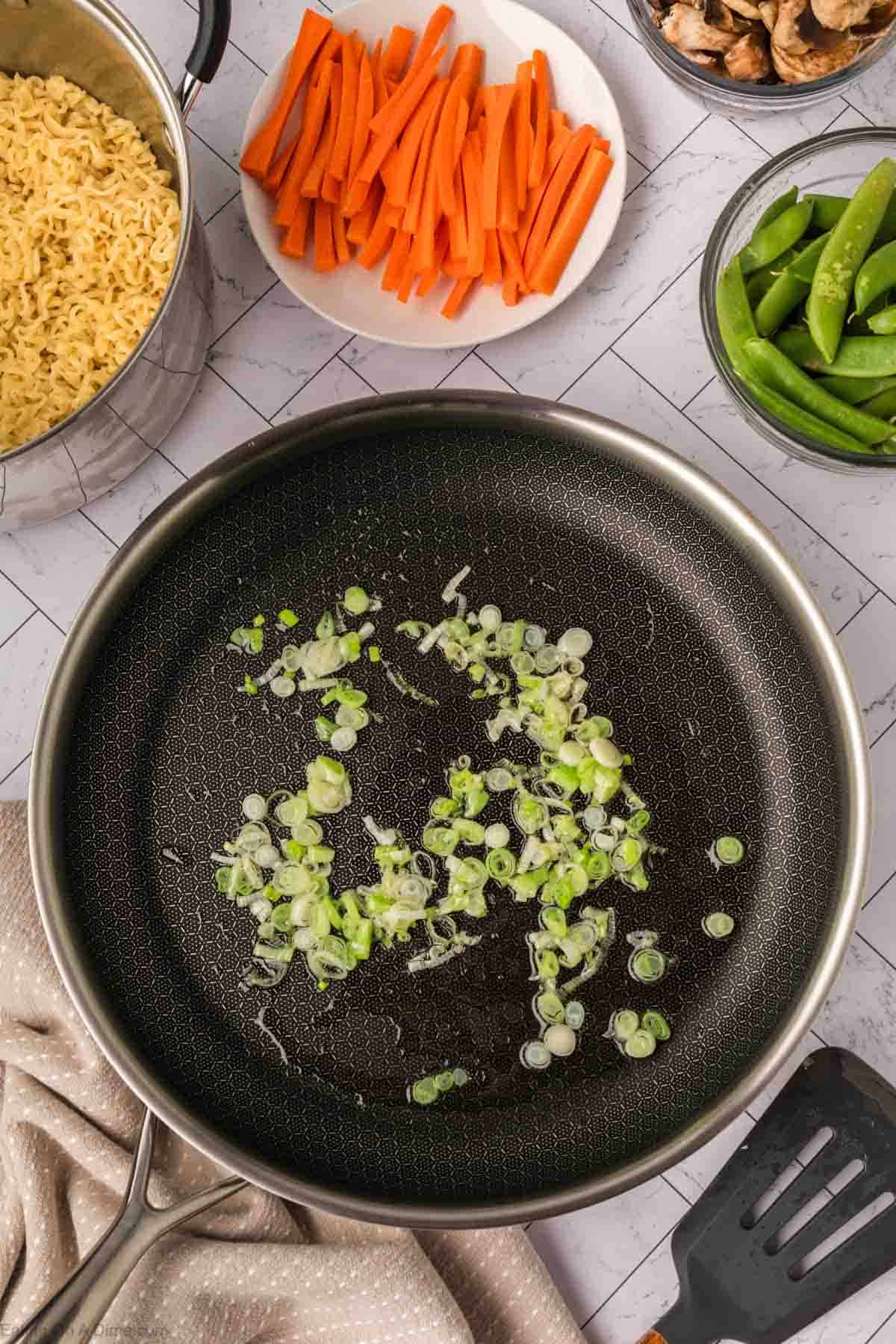 A frying pan with chopped green onions sautéing for a vibrant Ramen Stir Fry, surrounded by bowls of sliced carrots, snap peas, mushrooms, and a pot of cooked noodles on a white tiled surface.