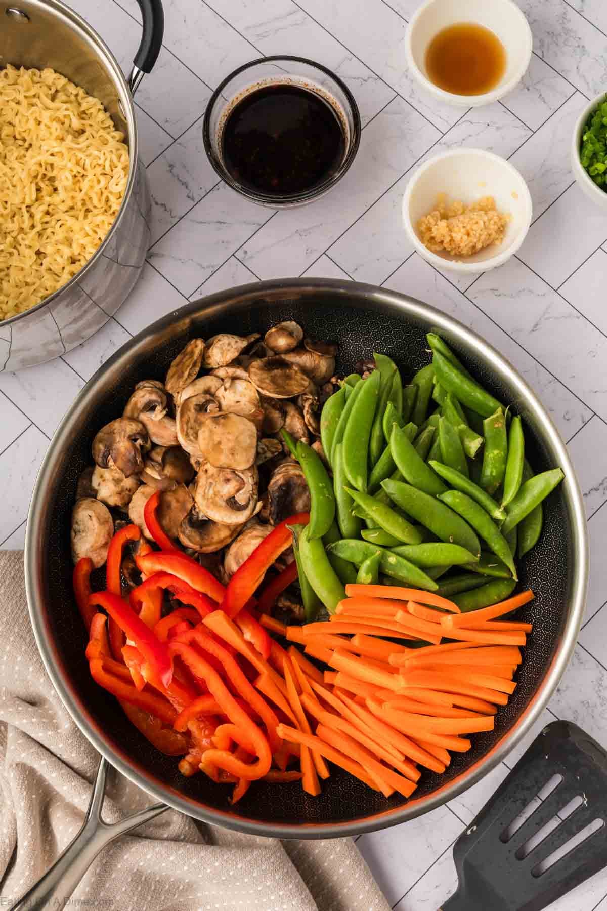 A frying pan with sliced mushrooms, snap peas, red bell peppers, and carrot sticks sits on a counter next to a pot of cooked noodles—perfect for creating a vibrant Ramen Stir Fry with bowls of sauce, garlic, and chopped green onions.