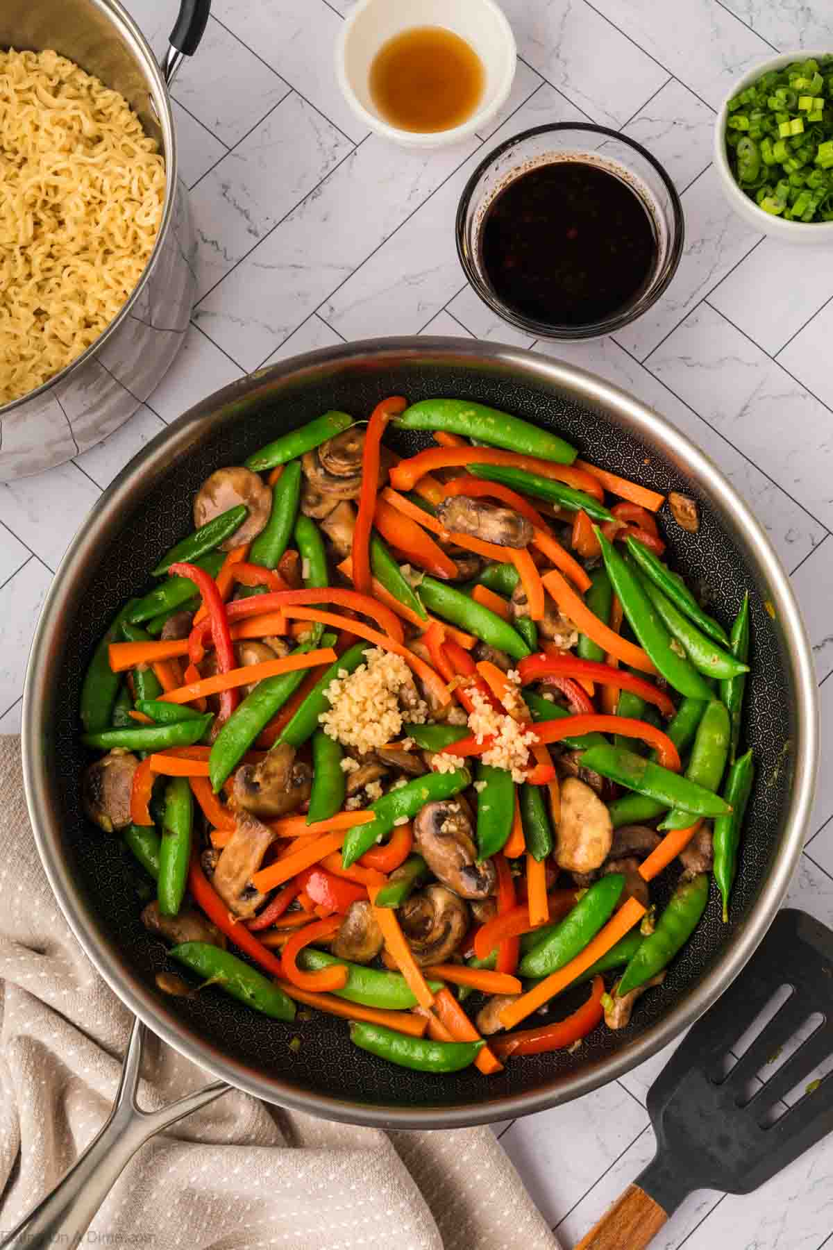 A skillet filled with snap peas, sliced red bell peppers, mushrooms, and minced garlic sits on a white counter—perfect for a colorful stir fry. Nearby are bowls of sauce, chopped green onions, cooked ramen noodles, and a spatula.