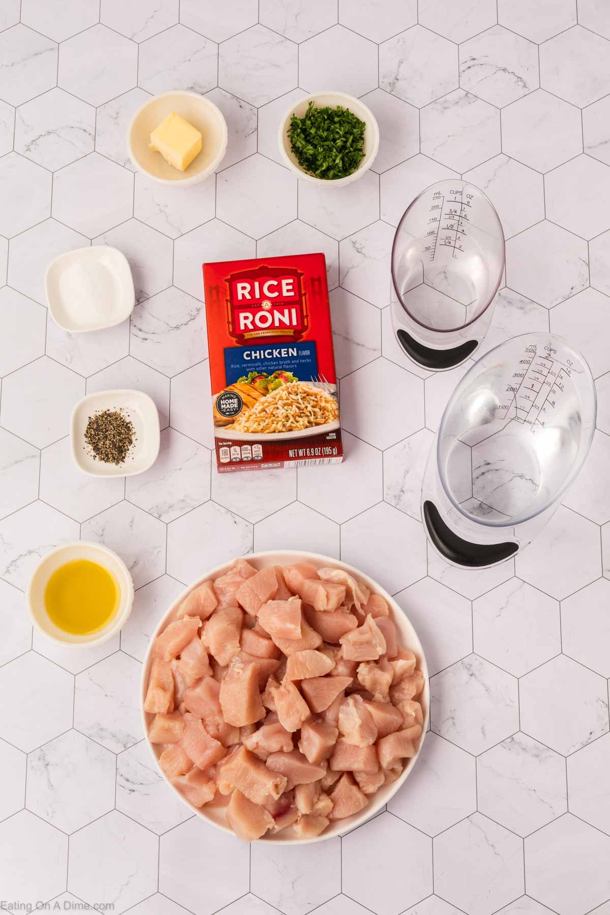 A bowl of diced raw chicken, a box of Rice A Roni Chicken, chopped herbs, butter, spices, oil, and two measuring cups are arranged on a white hexagonal tile surface for a tasty chicken casserole.