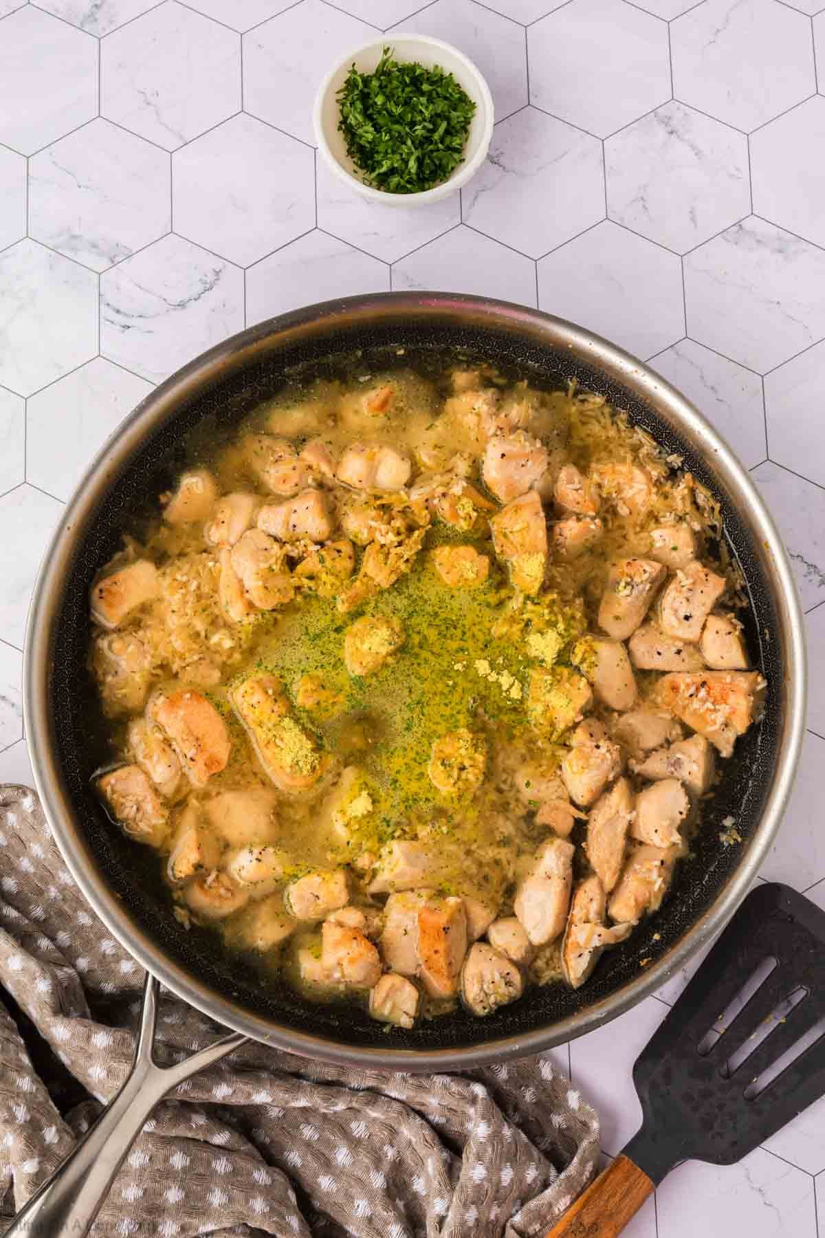 A skillet filled with cooked chicken pieces in a creamy Rice A Roni Chicken sauce, garnished with herbs. A small bowl of chopped green herbs sits above the skillet on a white, hexagon-patterned surface. A spatula and towel are nearby.