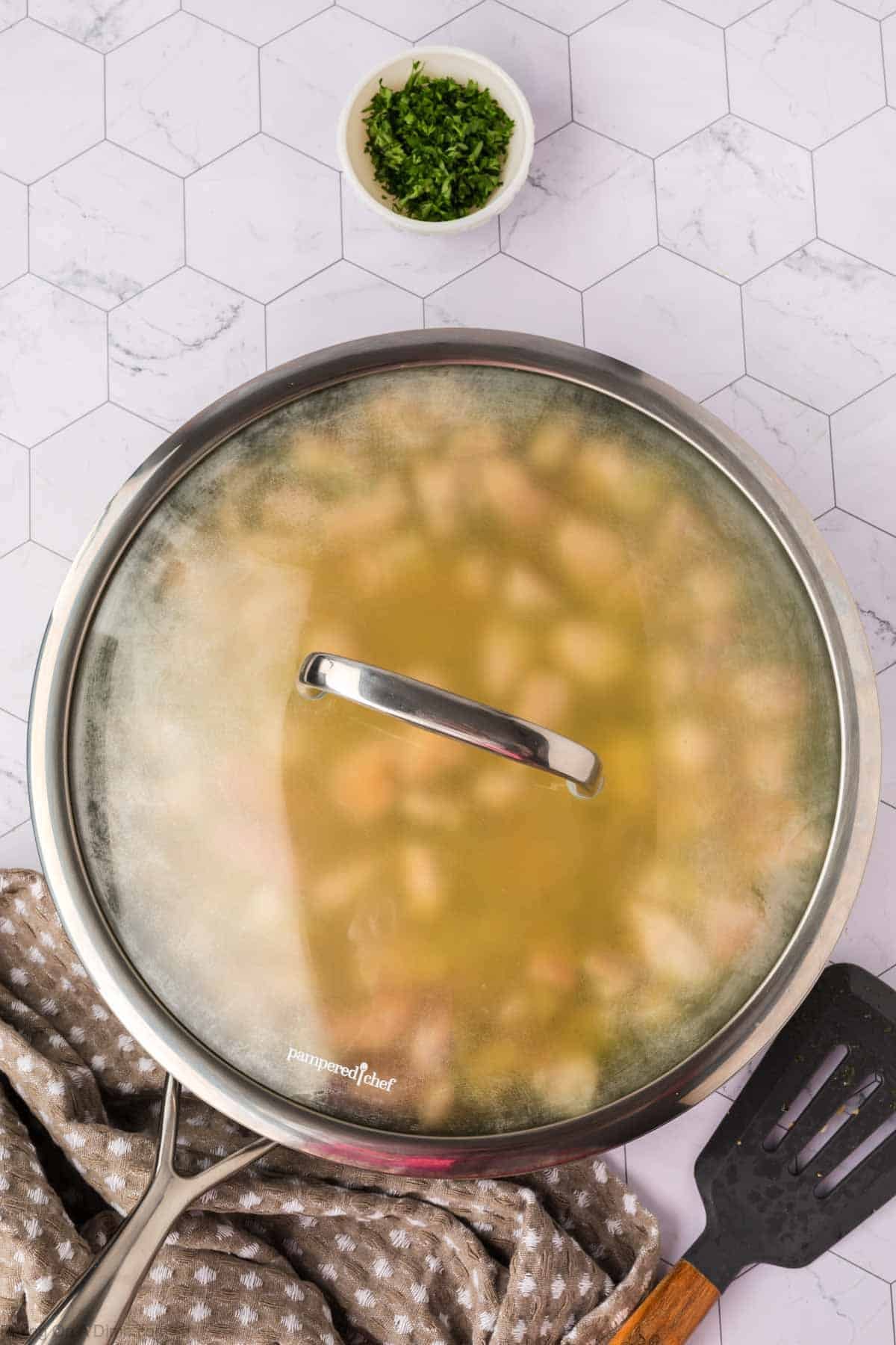 A large skillet with a glass lid steams on a marble surface, filled with a comforting chicken casserole. Nearby are a bowl of chopped herbs, a kitchen towel, and a black spatula.