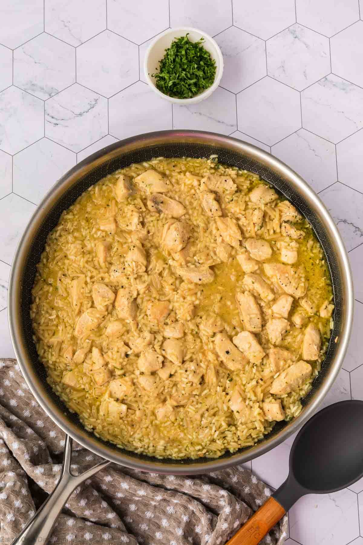 A skillet filled with creamy Rice A Roni Chicken and rice sits on a white hexagonal-tile surface, next to a bowl of chopped herbs, a gray spoon, and a brown-and-white towel.