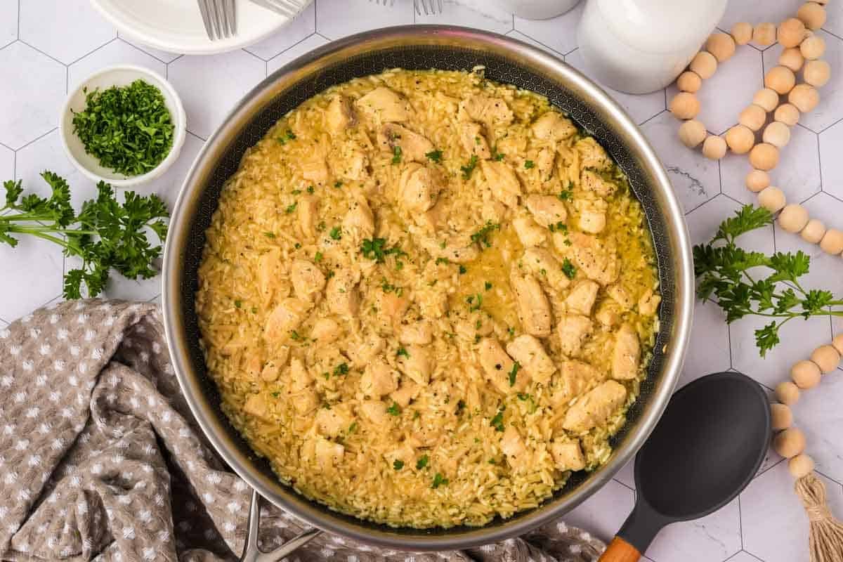A skillet filled with creamy Rice A Roni Chicken casserole, garnished with chopped parsley, sits on a white surface, surrounded by a bowl of fresh parsley, a serving spoon, plates, and a beige patterned cloth.