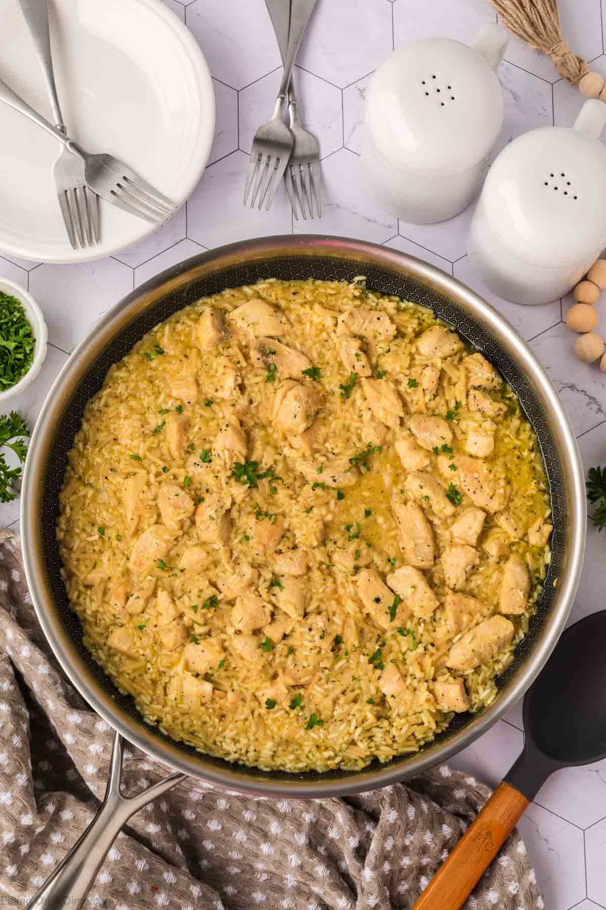 A skillet filled with creamy Rice A Roni Chicken casserole, garnished with chopped herbs, sits on a patterned cloth. Plates, forks, salt and pepper shakers, and small bowls with herbs surround the skillet on a white table.