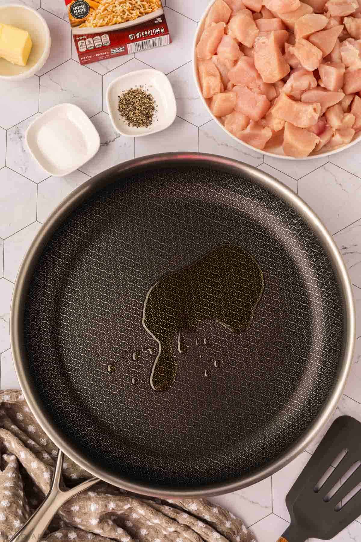 A frying pan with oil sits on a countertop next to diced raw chicken, a box of Rice A Roni Chicken, a pat of butter, small bowls of salt and pepper, and a spatula resting on a patterned towel.