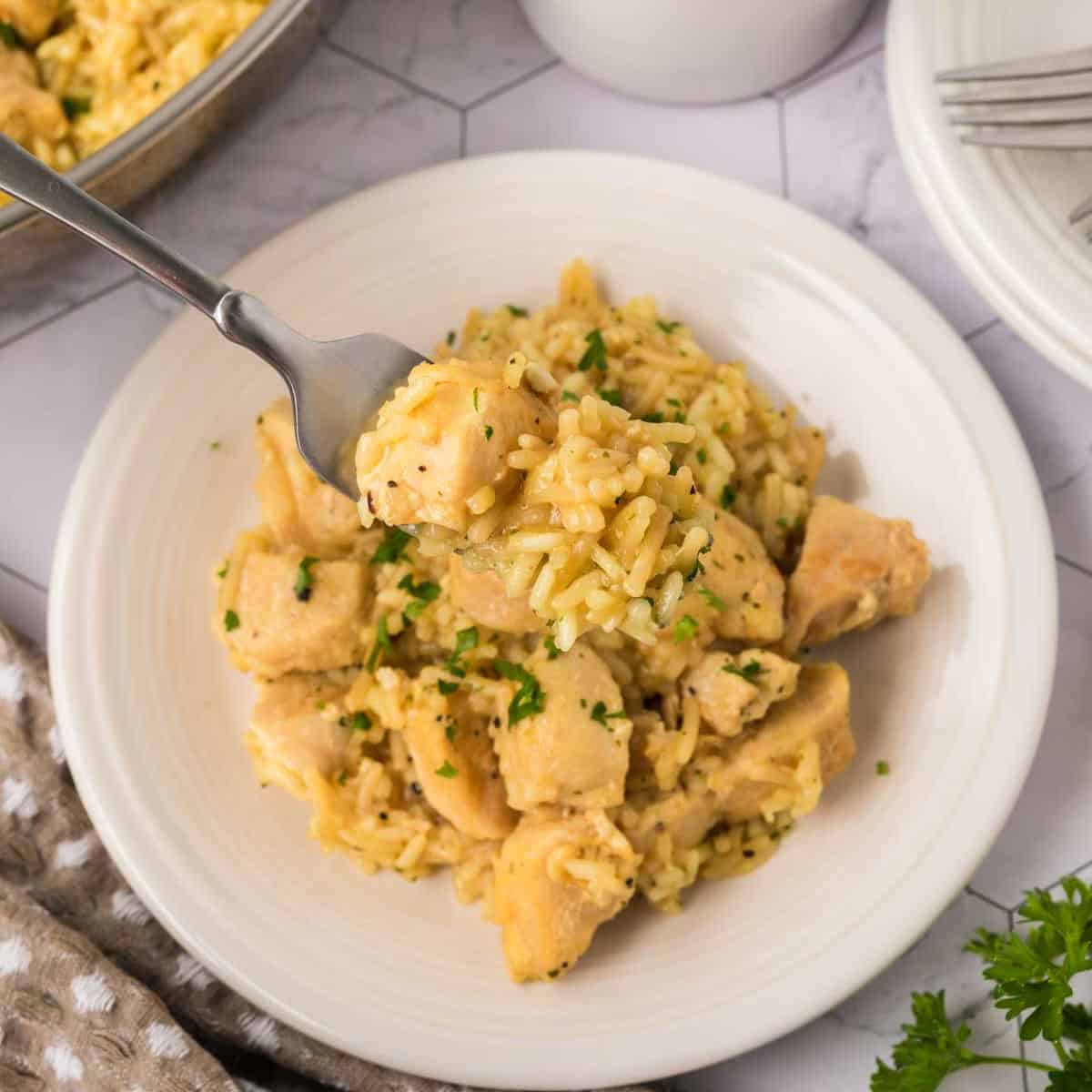 A white plate filled with creamy Rice A Roni Chicken casserole and chunks of cooked chicken, garnished with chopped herbs. A fork lifts a bite-sized portion above the plate. Other dishes and utensils are visible in the background.