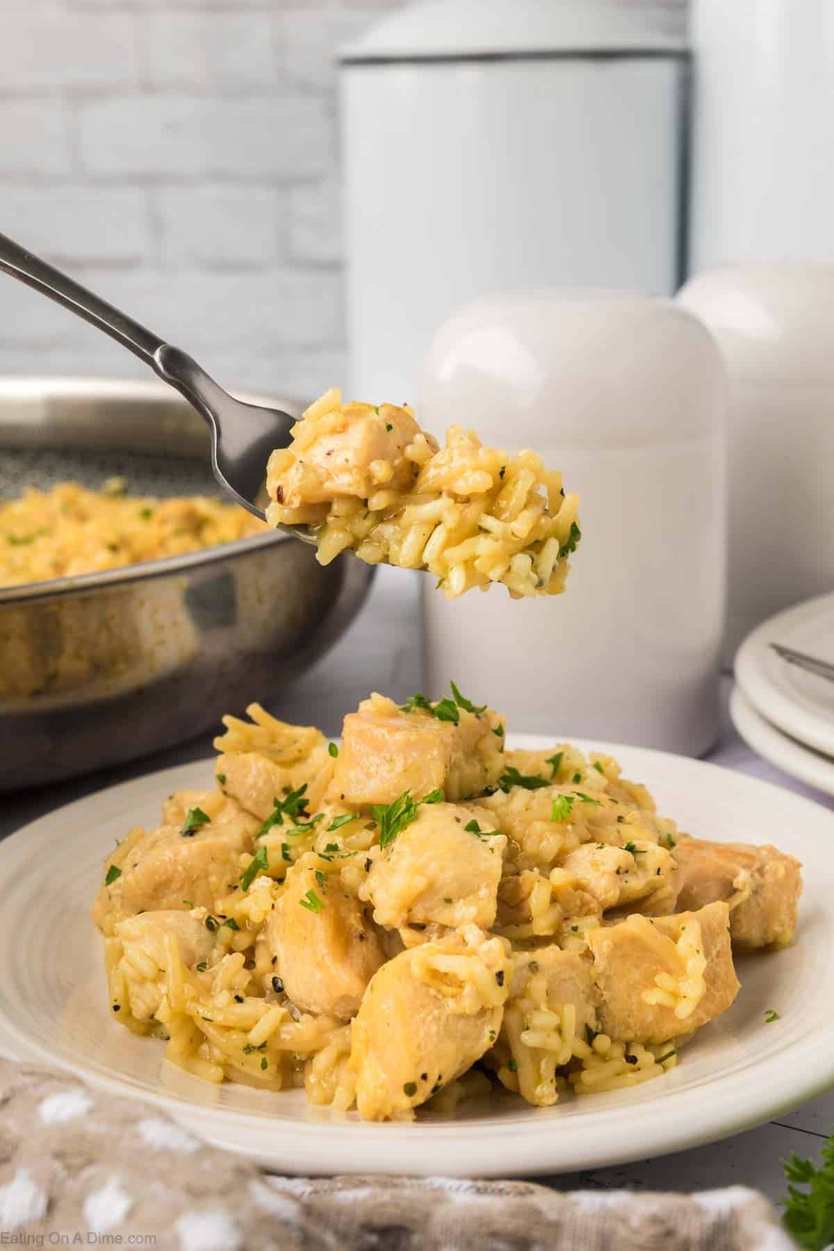 A close-up of a plate of creamy Rice A Roni Chicken casserole garnished with parsley. A fork holds a bite above the plate. In the background, a pan of the same dish and some white kitchen containers are visible.