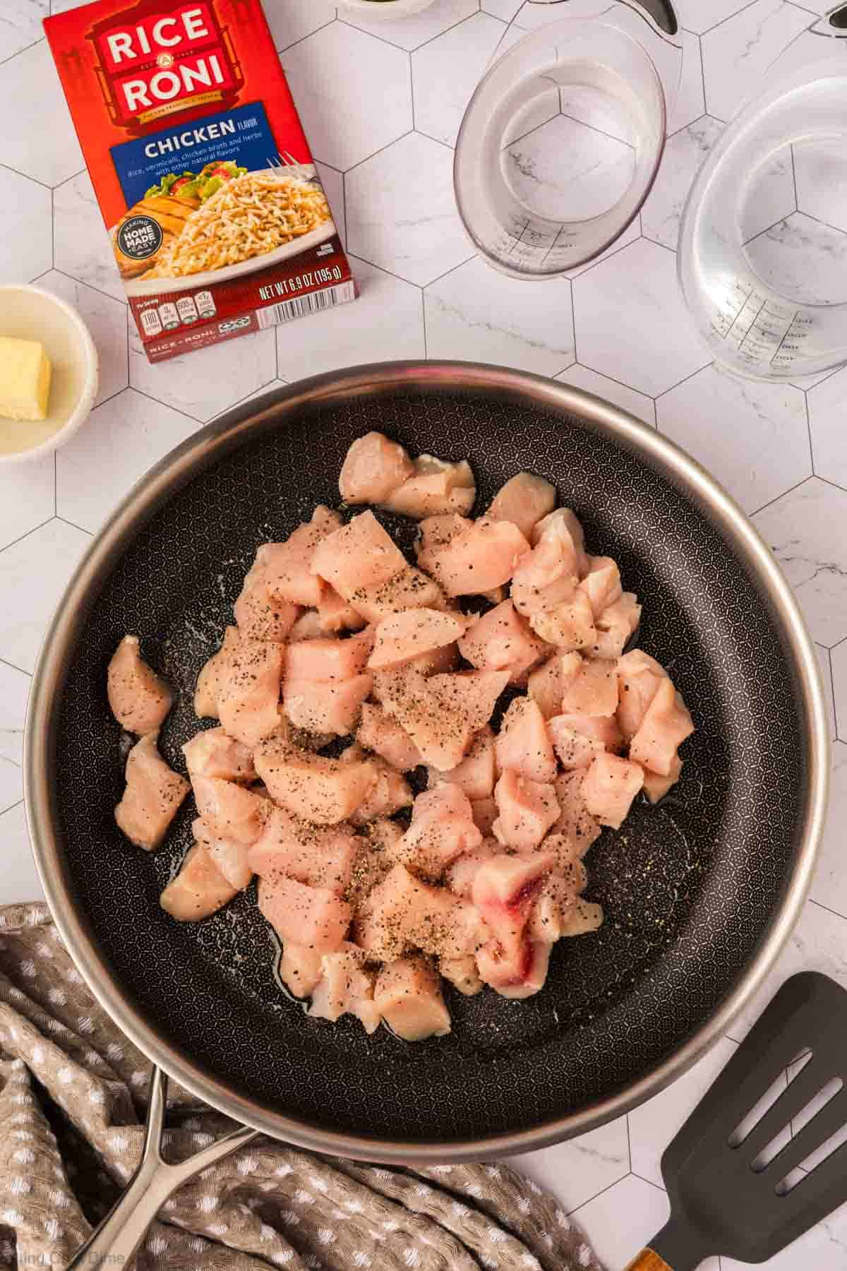 A frying pan with raw, diced chicken seasoned with pepper, a box of Rice A Roni Chicken flavor, two measuring cups with water, a pat of butter, a spatula, and a patterned cloth—perfect for starting an easy Rice A Roni Chicken casserole.