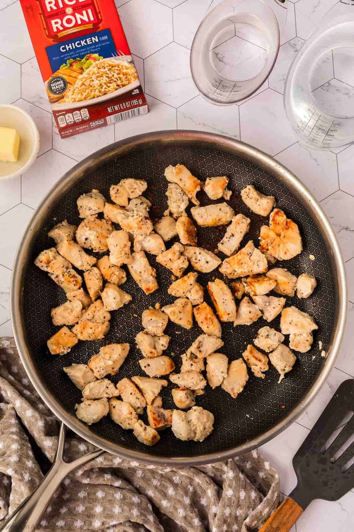 A skillet with cooked, diced chicken pieces sits on a towel. Nearby are a box of Rice A Roni Chicken, a measuring cup of water, a pat of butter, and a black spatula on a white hexagon-patterned surface—perfect for chicken casserole prep.