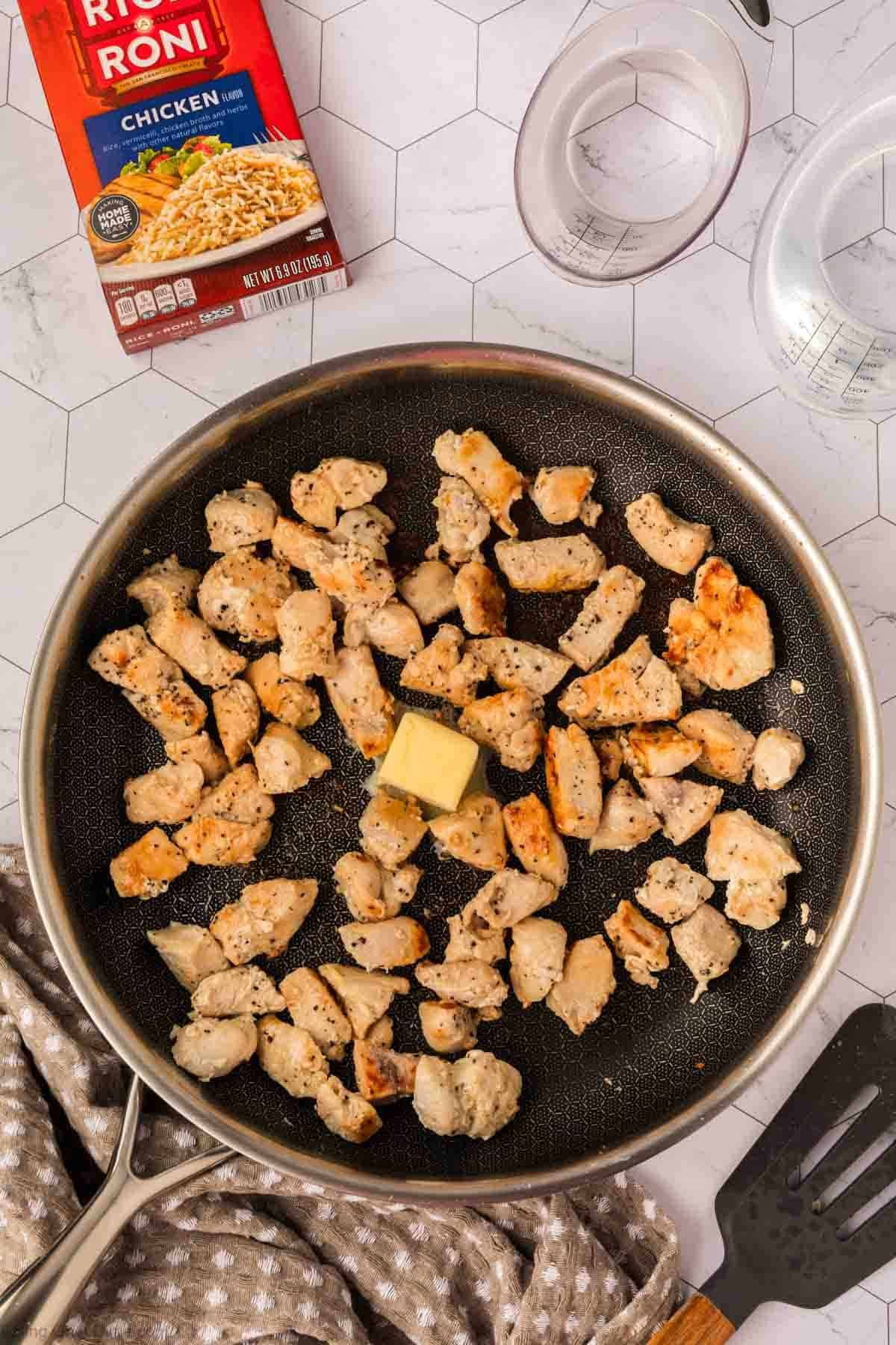 A frying pan with cooked, seasoned chicken pieces and a pat of butter in the center. Nearby are a Rice A Roni Chicken flavor box, two plastic measuring cups, a spatula, and a beige kitchen towel with white dots.