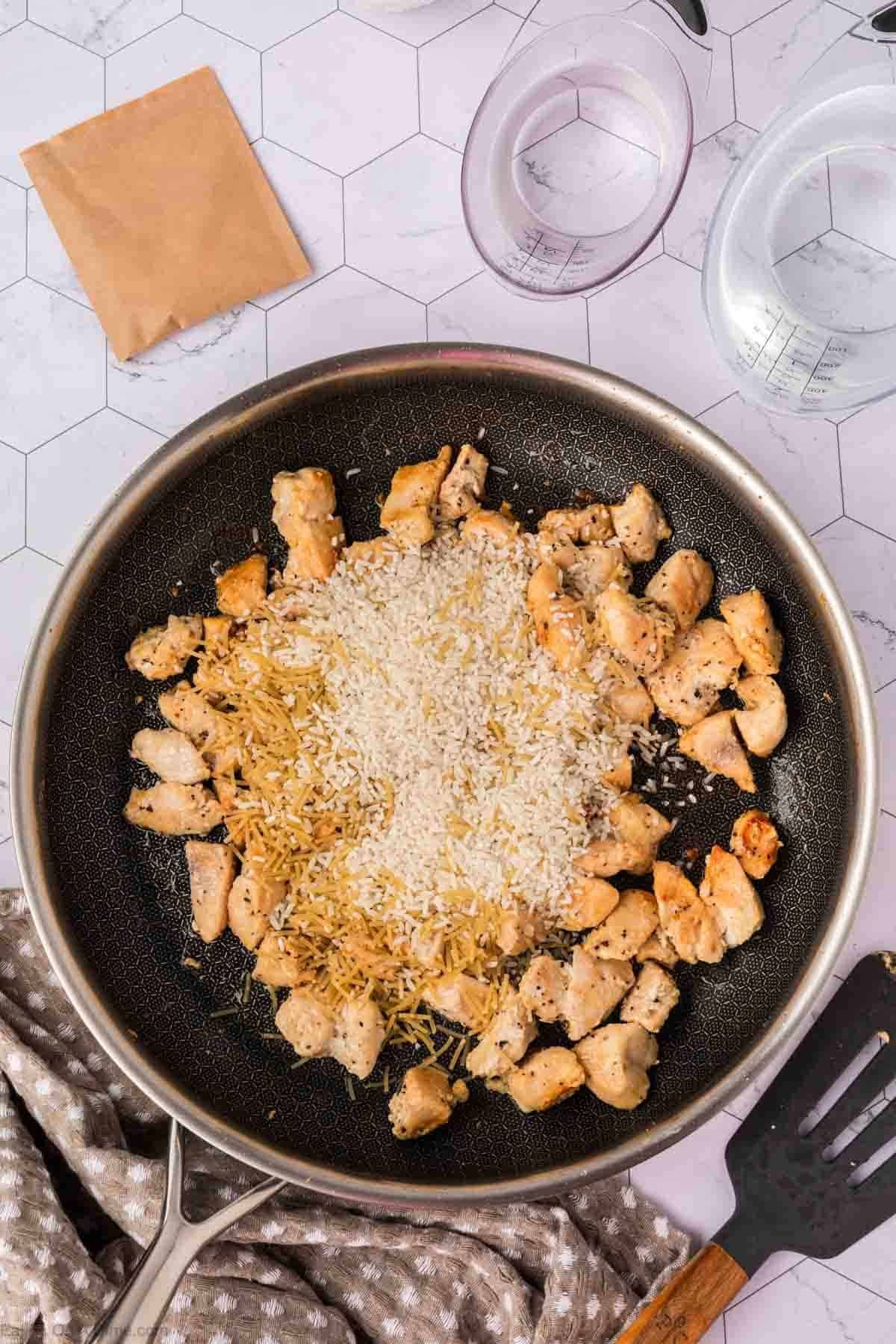 A frying pan contains cooked chicken pieces, uncooked Rice A Roni Chicken, and pasta on a stovetop. Nearby are a spatula, measuring cups, a brown packet, and a textured towel on a hexagon-patterned surface.