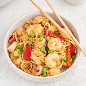 A bowl of shrimp lo mein with noodles, red bell peppers, green onions, and sesame seeds, served with chopsticks on a light surface—perfect for adding to your Meal Plan 87.