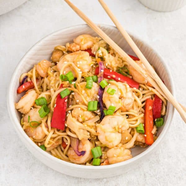 A bowl of shrimp lo mein with noodles, red bell peppers, green onions, and sesame seeds, served with chopsticks on a light surface—perfect for adding to your Meal Plan 87.